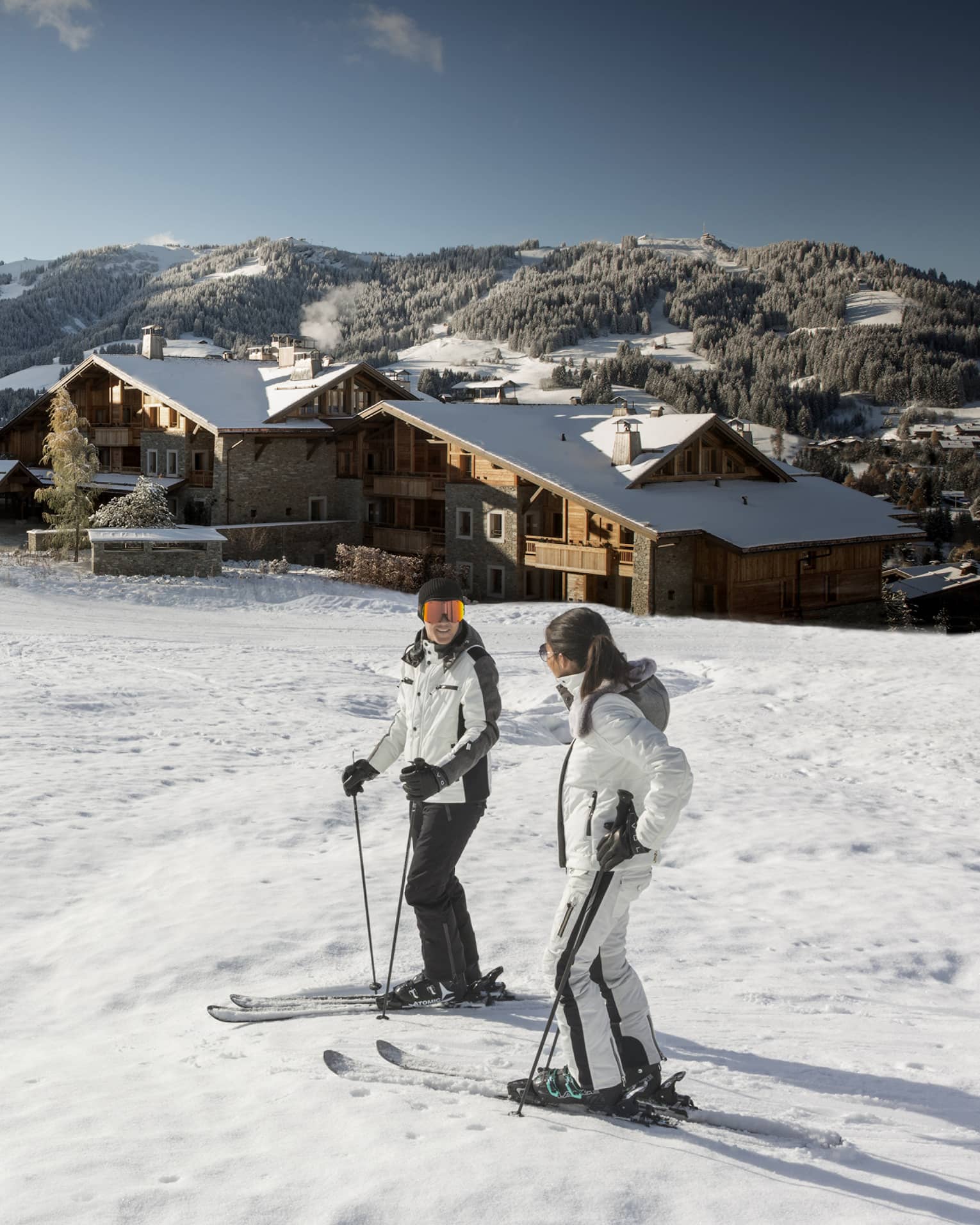 Man, woman on skis at top of snowy hill, Four Seasons Hotel Megeve in background