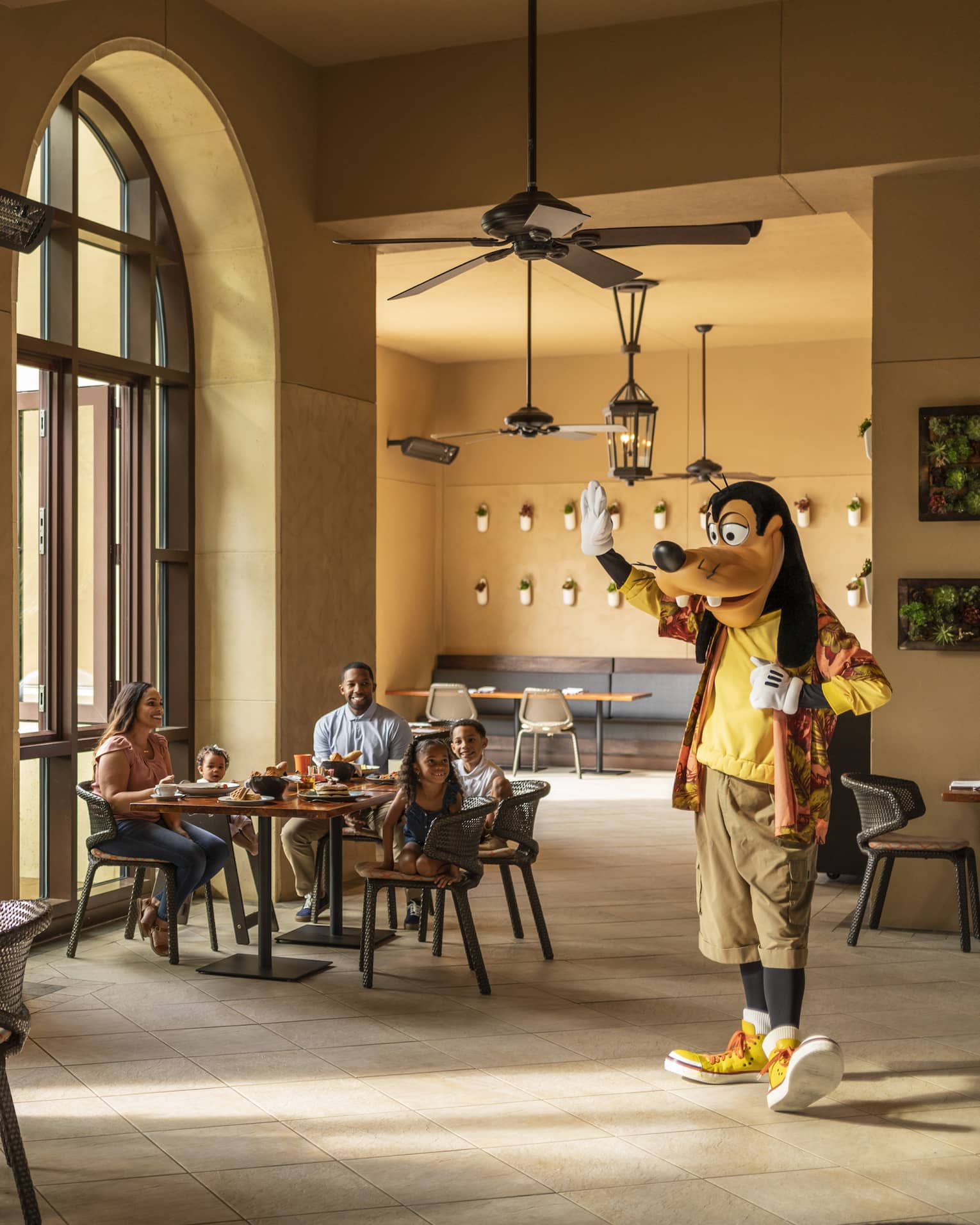 Sunlight filters into a restaurant where two families with young children smile at Goofy as he walks over and waves at them.
