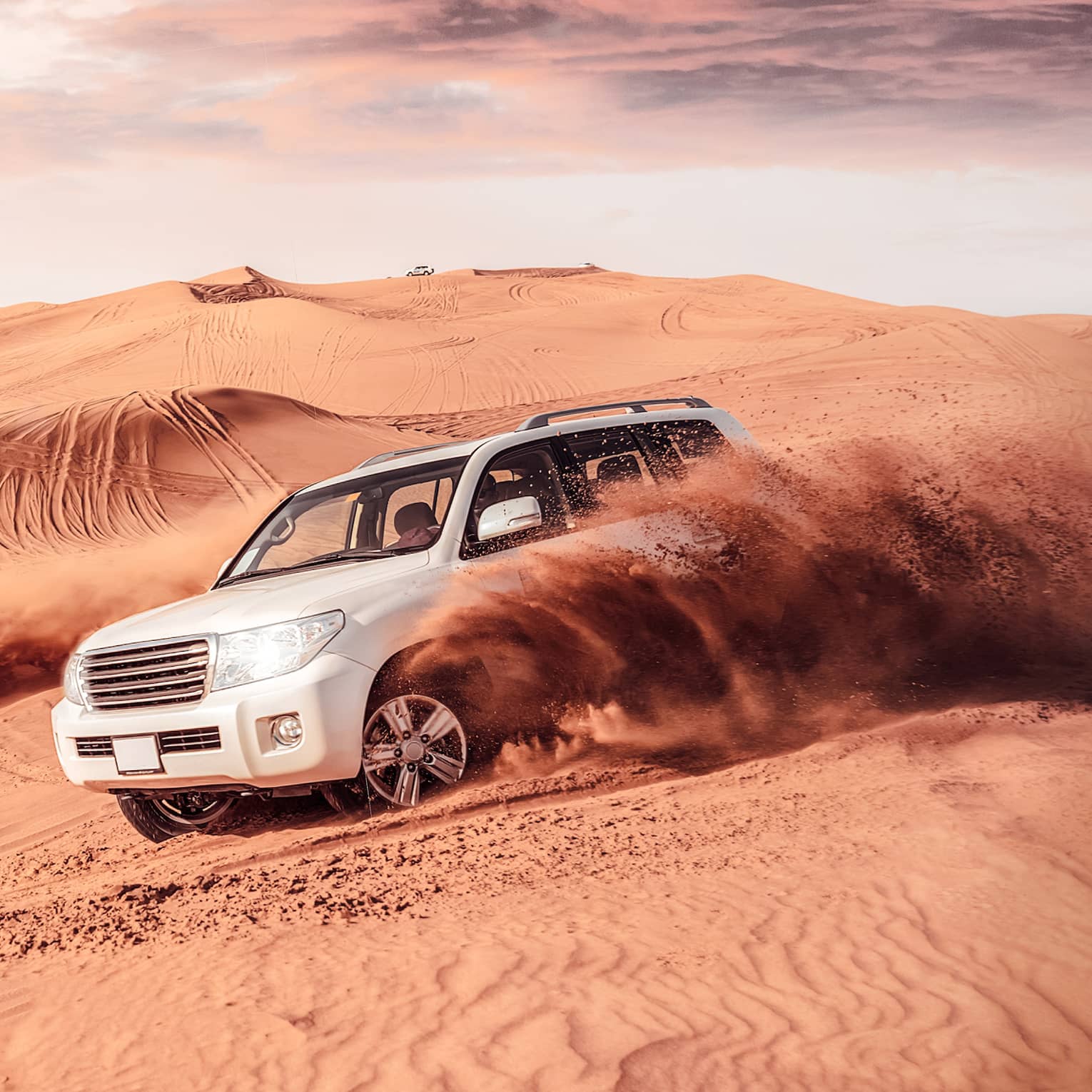 Sand flies from tires of two white SUV's driving across dunes
