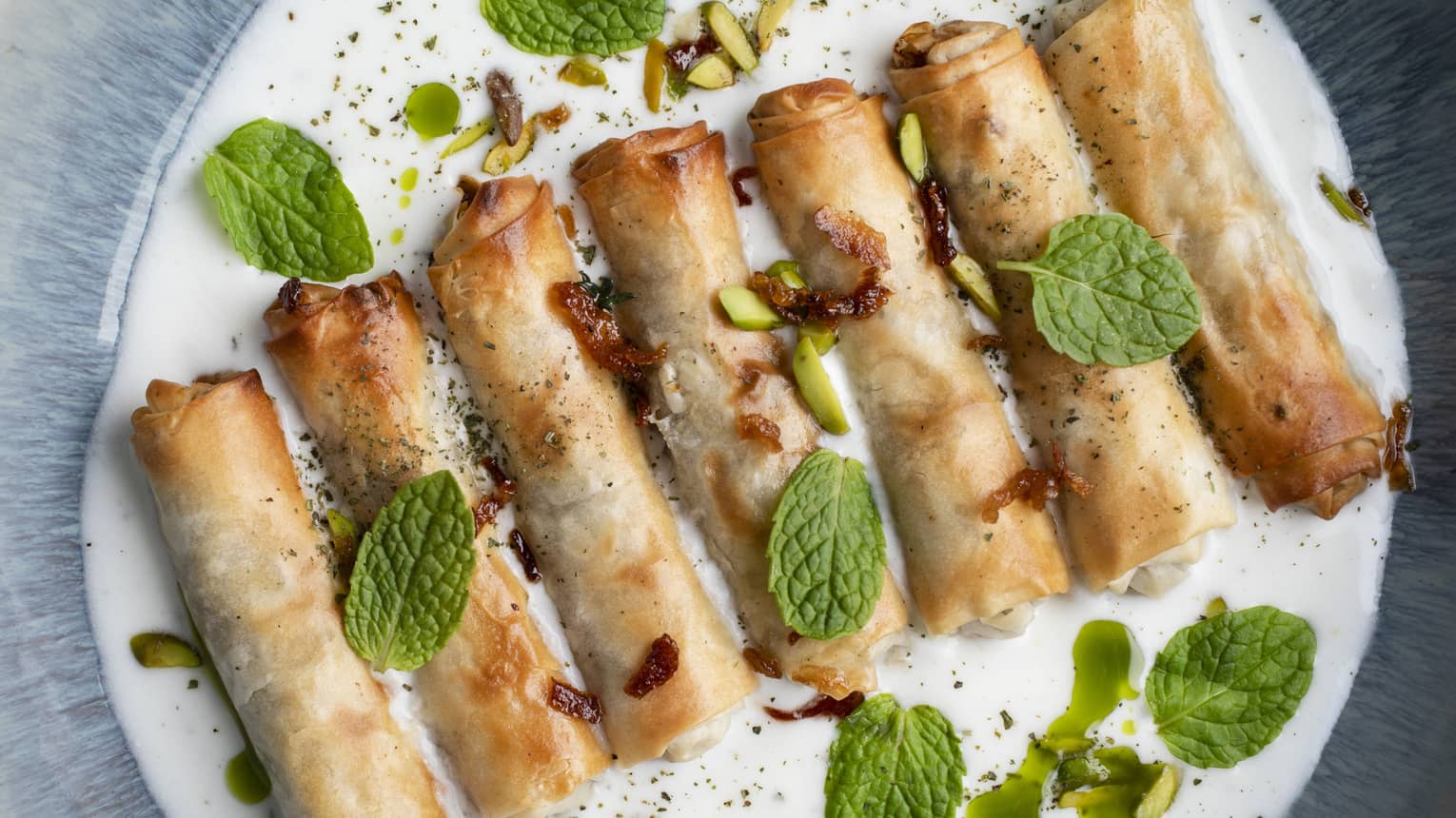 Seven baked meat rolls served in a row with mixed nuts, yogurt mint sauce and mint leaves as garnish