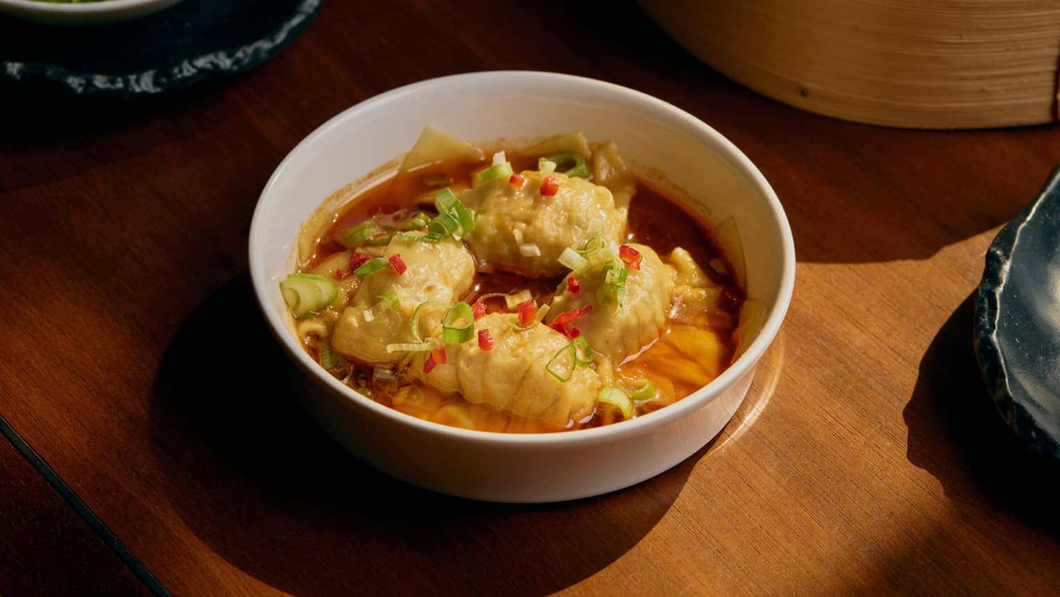 A wood table with several plates of food. One white bowl contains wontons with garnishes.