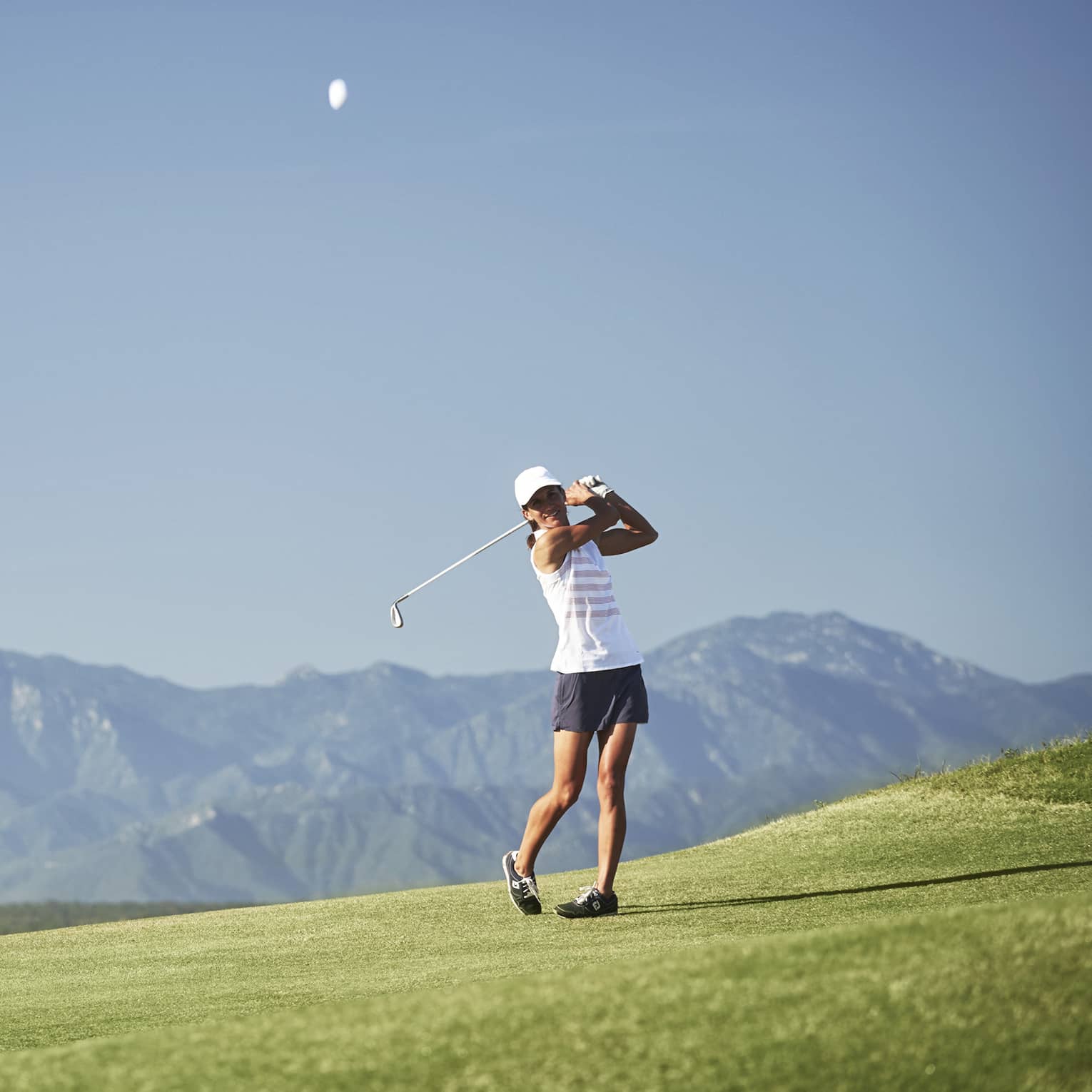On a smooth fairway against a striking panorama of mountains, a golfer ends their swing with a confident follow-through.
