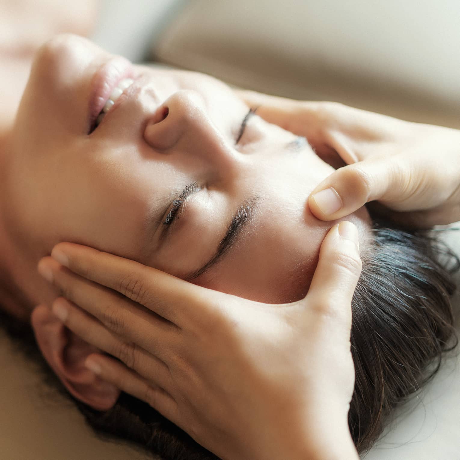 Spa facial, two hands rest on woman's forehead as she closes her eyes, lays under sheet on treatment table