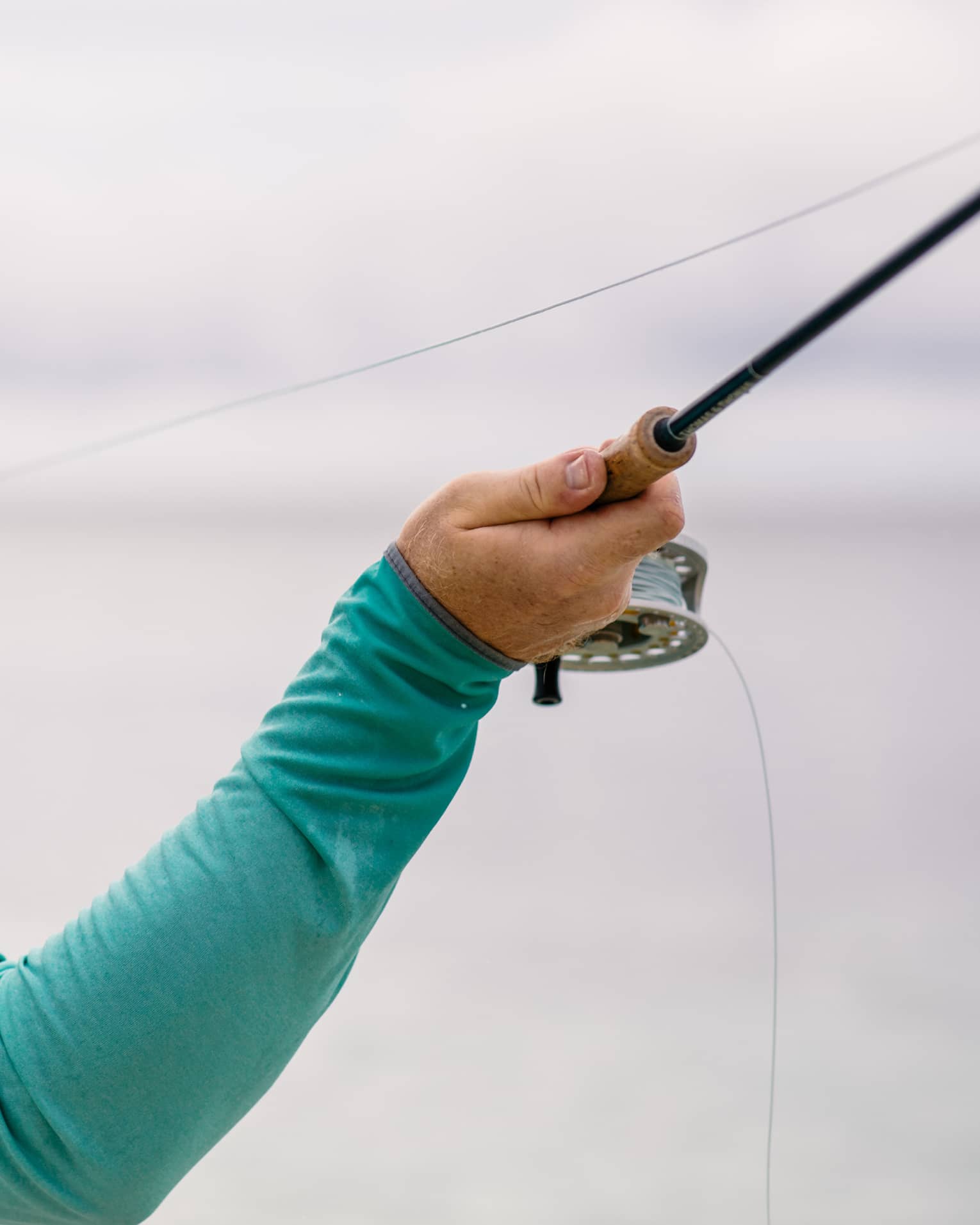 A fisherman facing away holds his fishing line in his left hand and draws back his rod with his right, readying to cast.