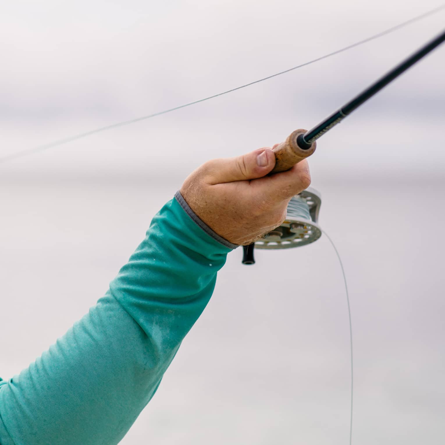 A fisherman facing away holds his fishing line in his left hand and draws back his rod with his right, readying to cast. 