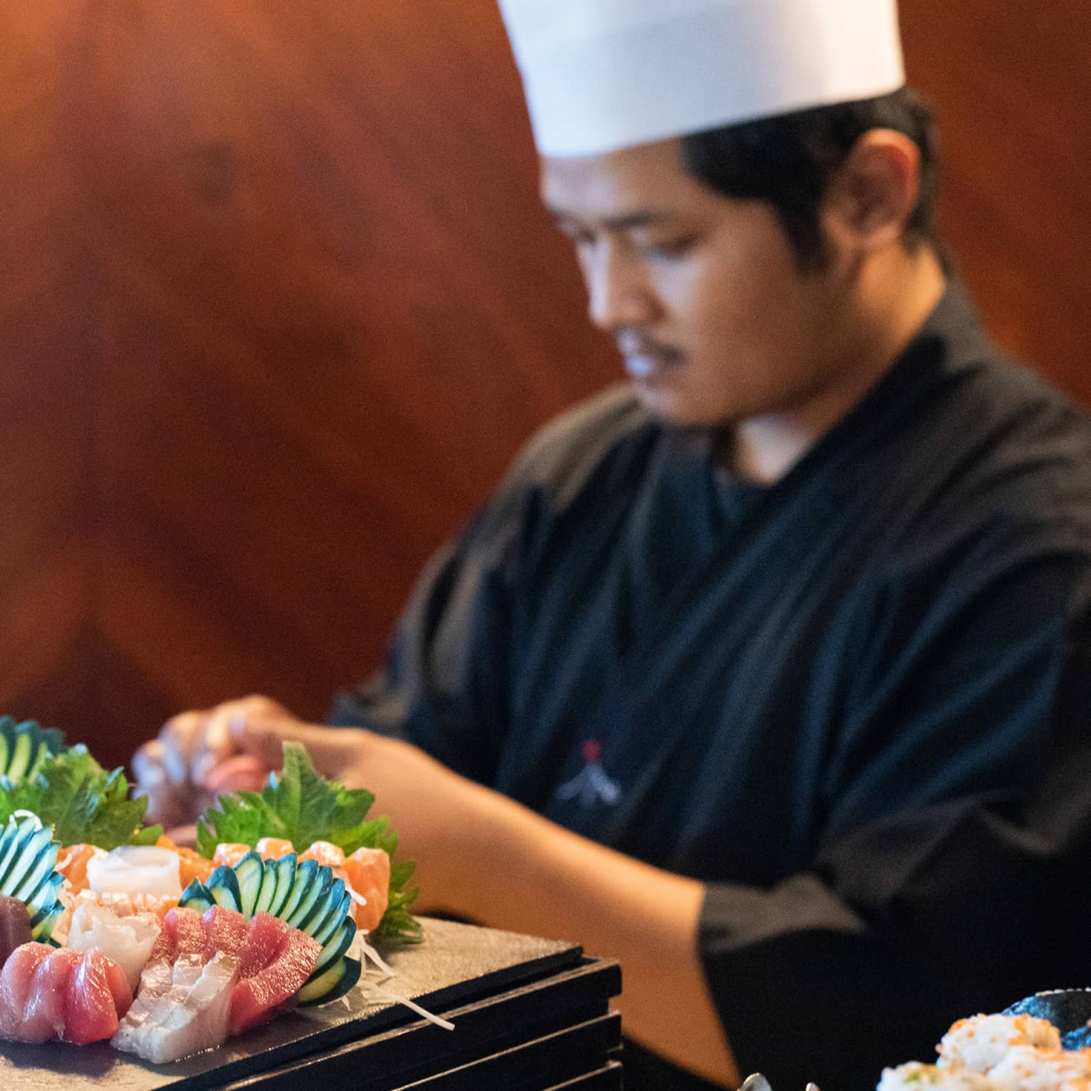 A cook in a chef's hat prepares an opulent sushi spread with decorative fans of cucumber slices on a thick wood platform.