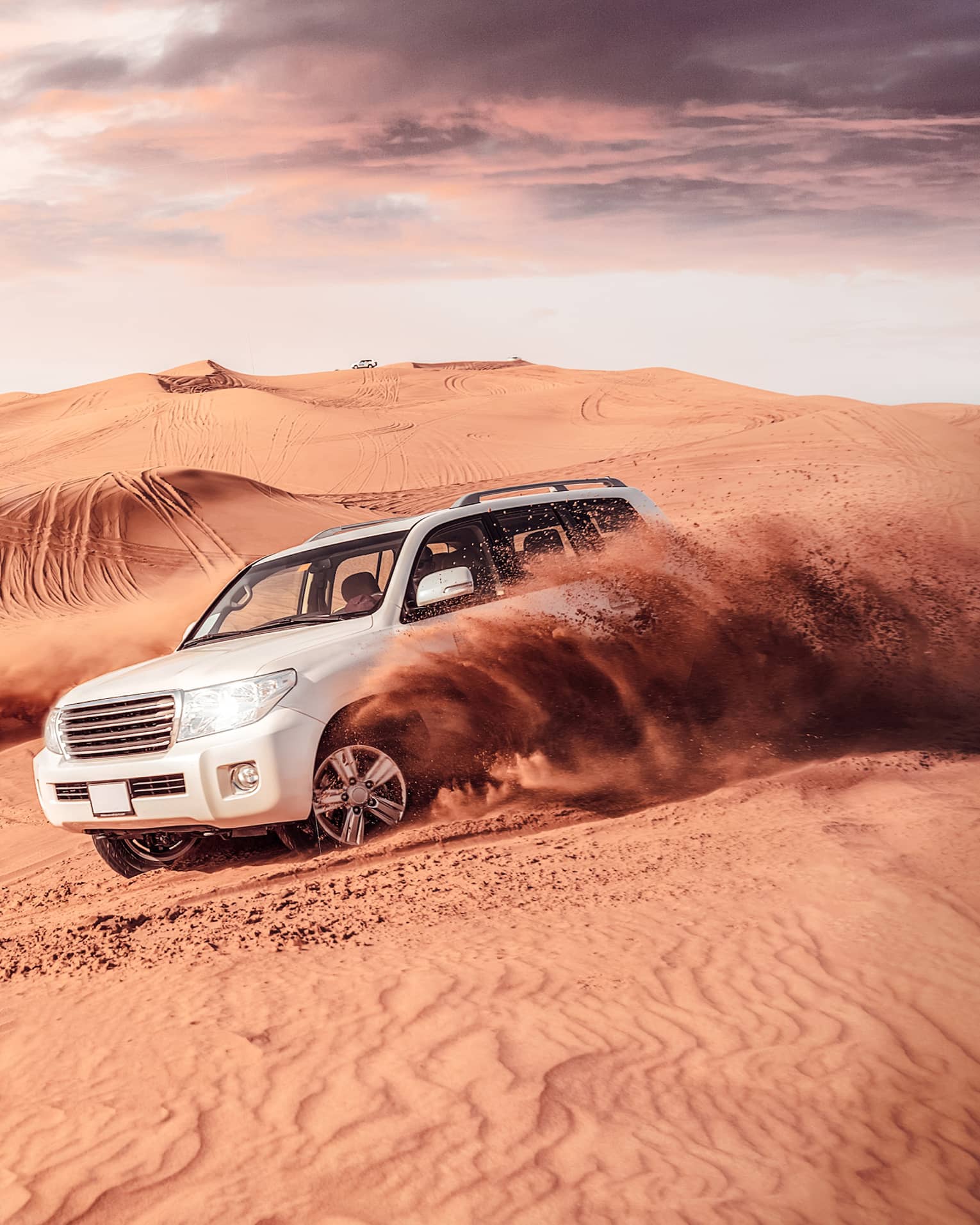 Sand flies from tires of two white SUV's driving across dunes