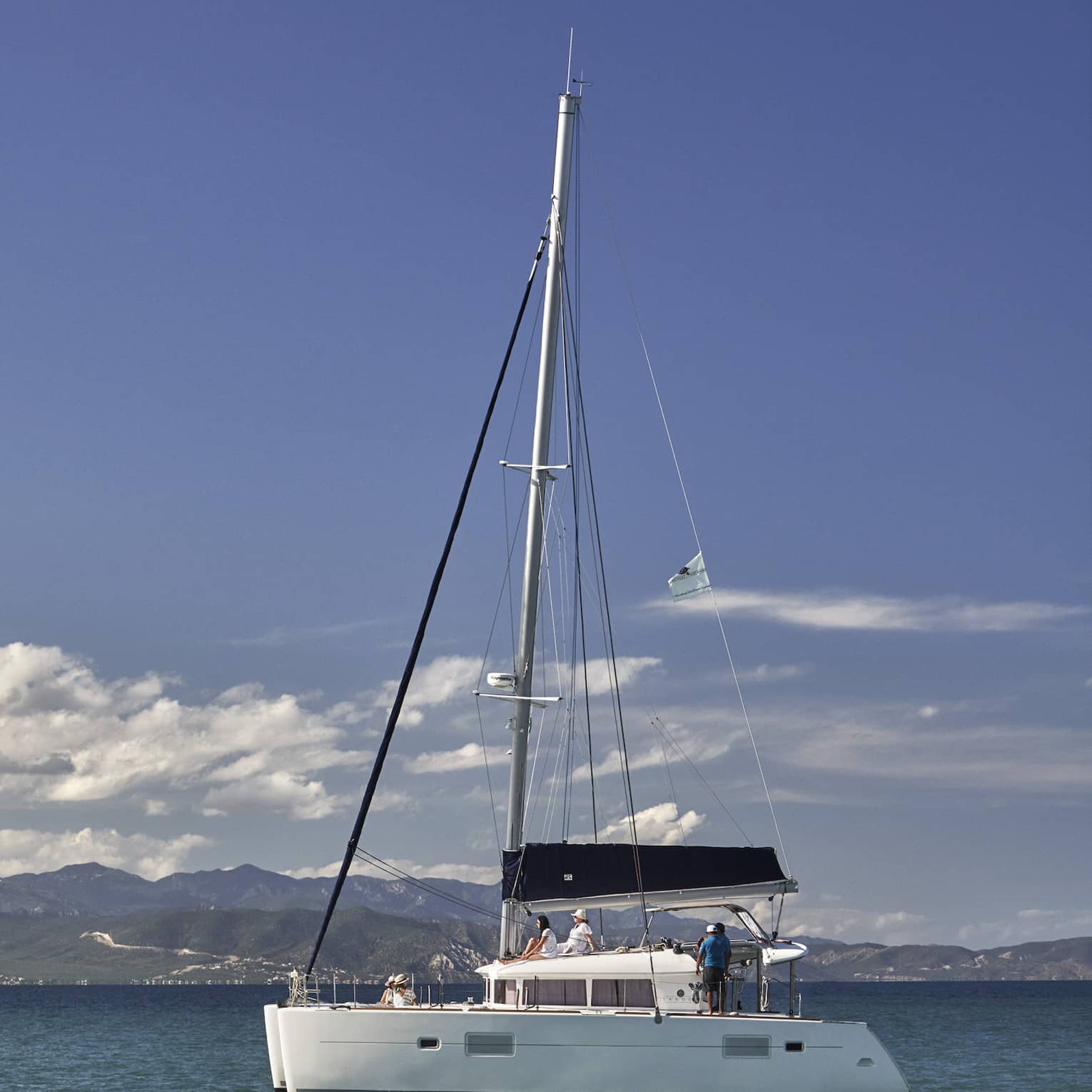 A catamaran on calm, blue water under a partly-cloudy sky with low green hills in the background and mountain ranges beyond.