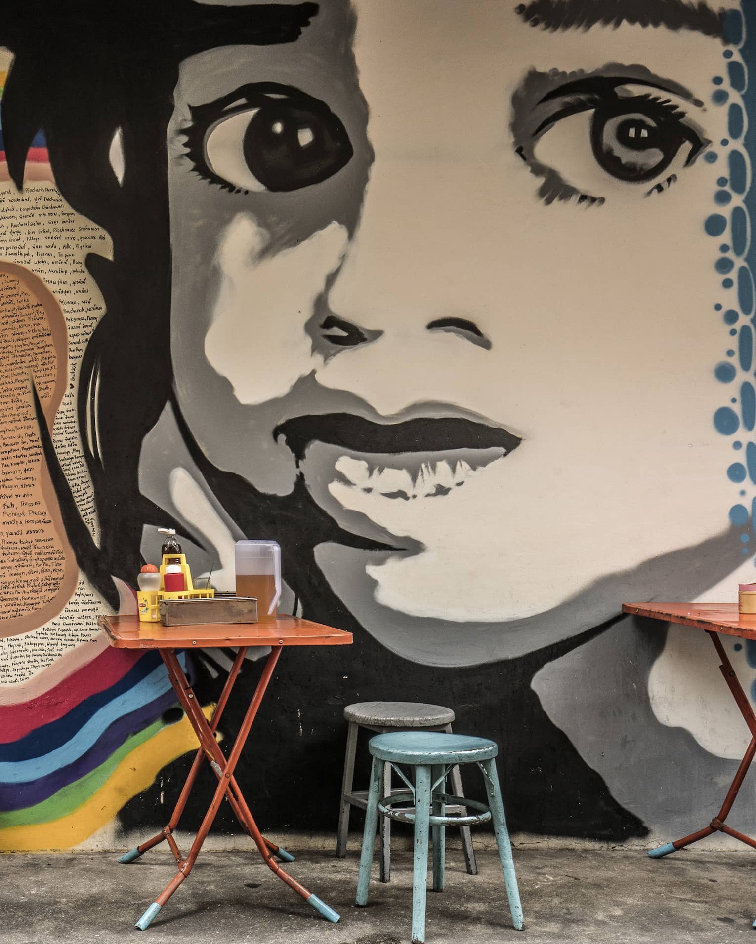 Street art mural featuring a large black-and-white face on a wall, with small, colourful dining tables and stools in front it