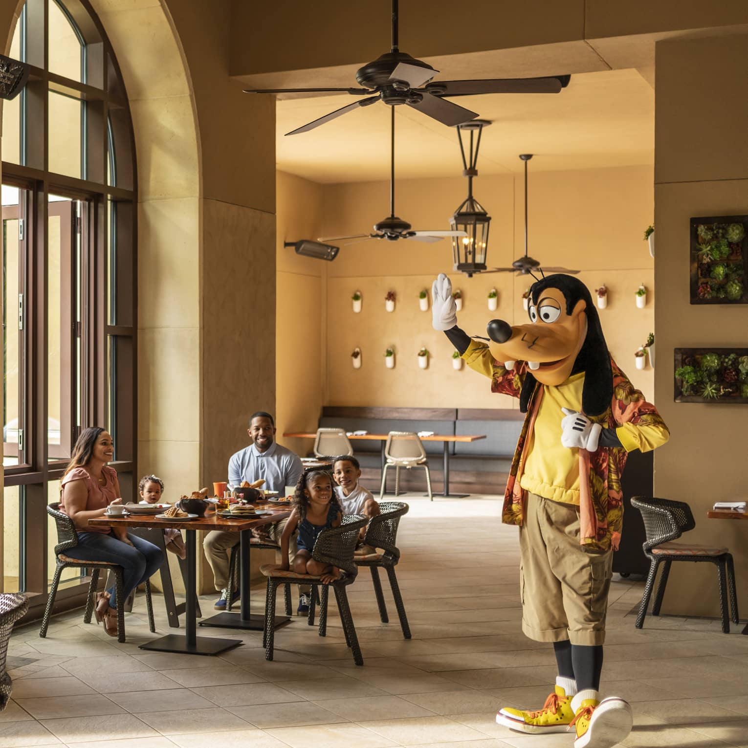Sunlight filters into a restaurant where two families with young children smile at Goofy as he walks over and waves at them.