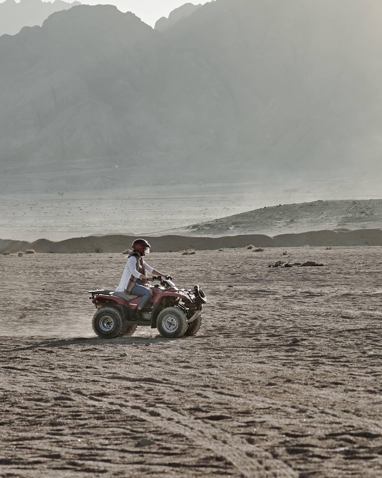A person in a helmet rides an ATV over an arid, dusty landscape past large hazy mountains that fill most of the image.