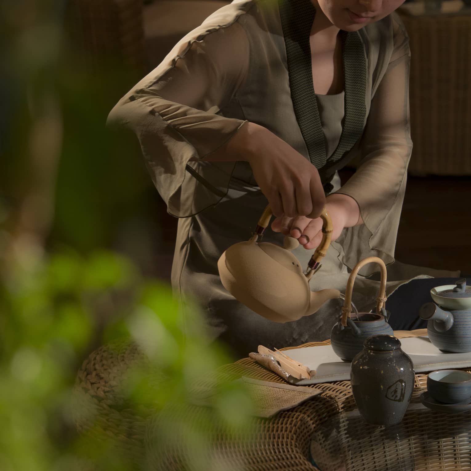 A server sits on an ottoman and pours from a stoneware kettle into a small pot that sits on a low wicker and glass table.
