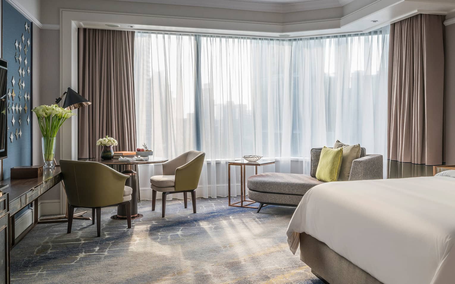 Sunny Deluxe Room bed with white linens, small dining table and chaise lounge, white curtains
