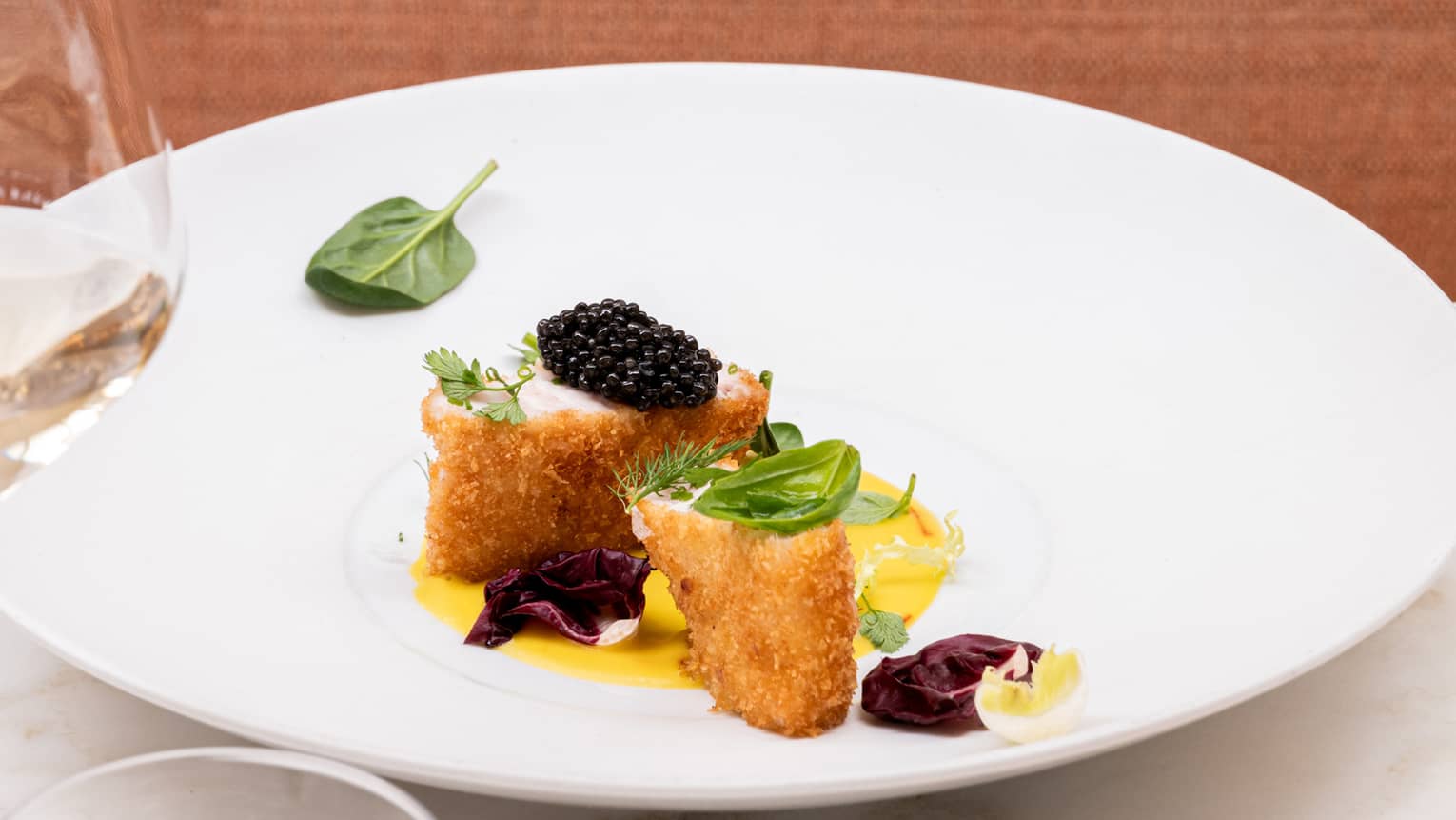 Breaded fish steaks garnished with black caviar and fresh herbs atop a golden puree in a wide dish, served with white wine.
