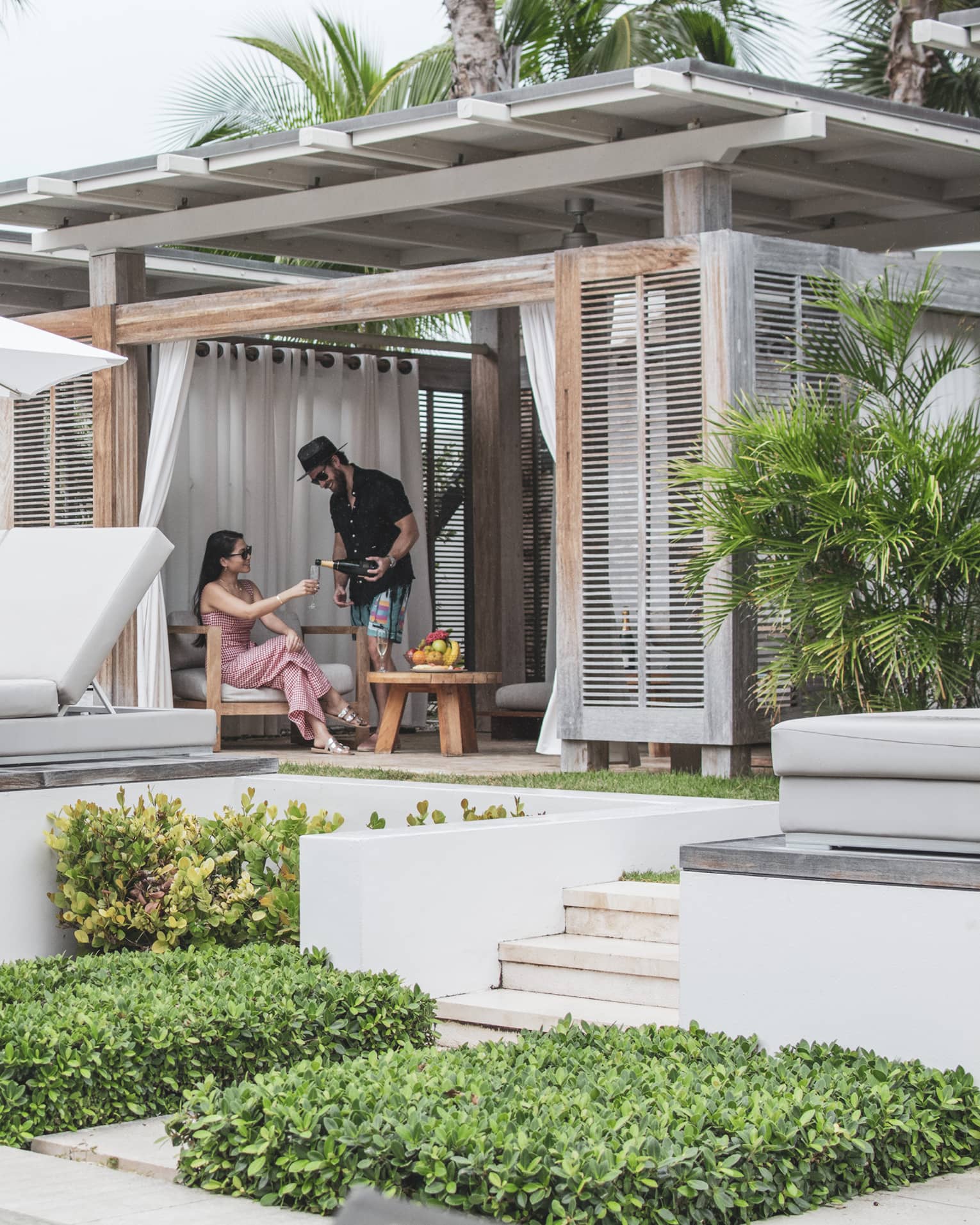 Two talk to each other in a covered pool cabana with a row of white daybeds and a row of grey lounge chairs and white umbrellas in front of them