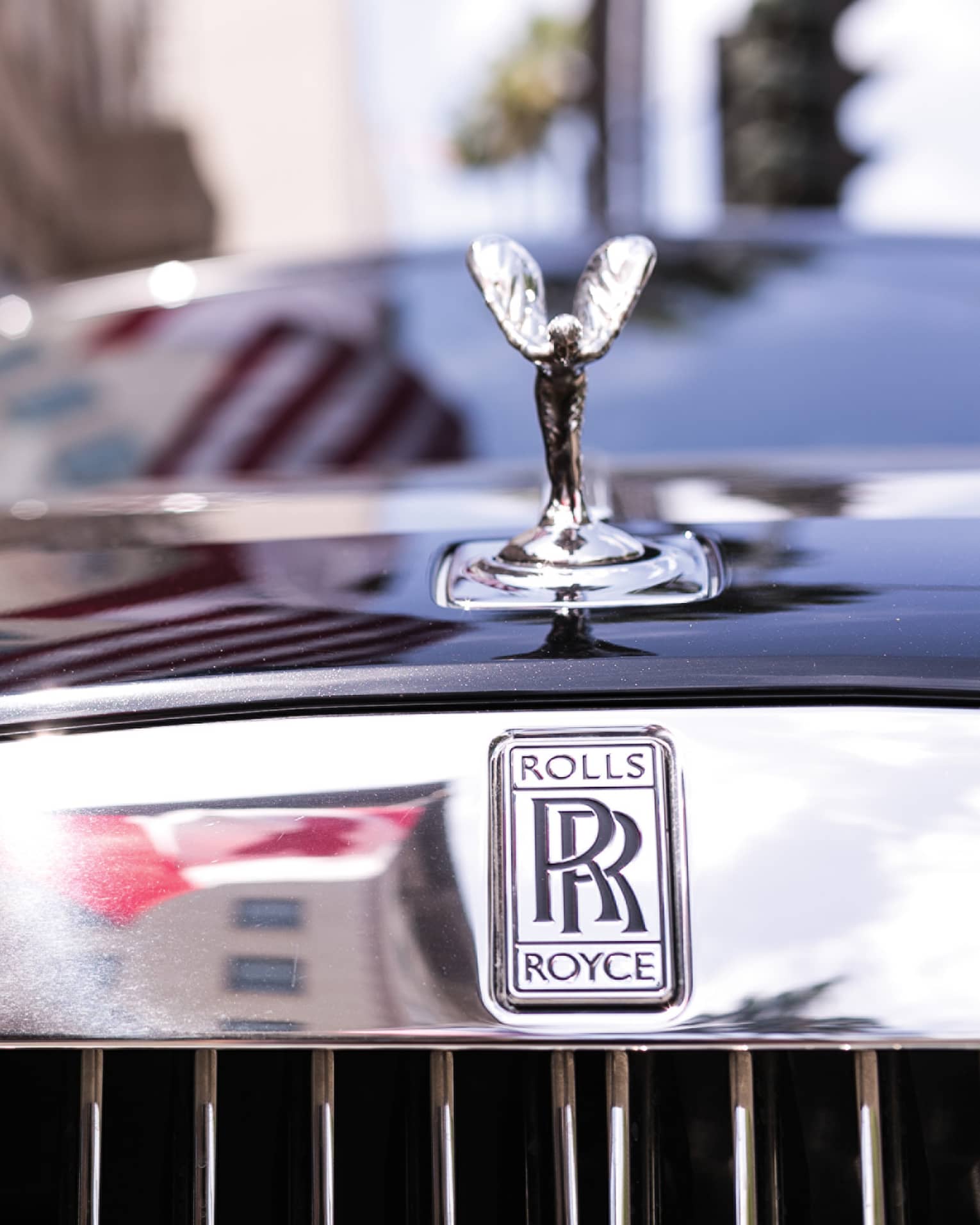 Close-up view of a winged silver ornament on the hood of black Rolls Royce, double-R logo on reflective chrome above grille.