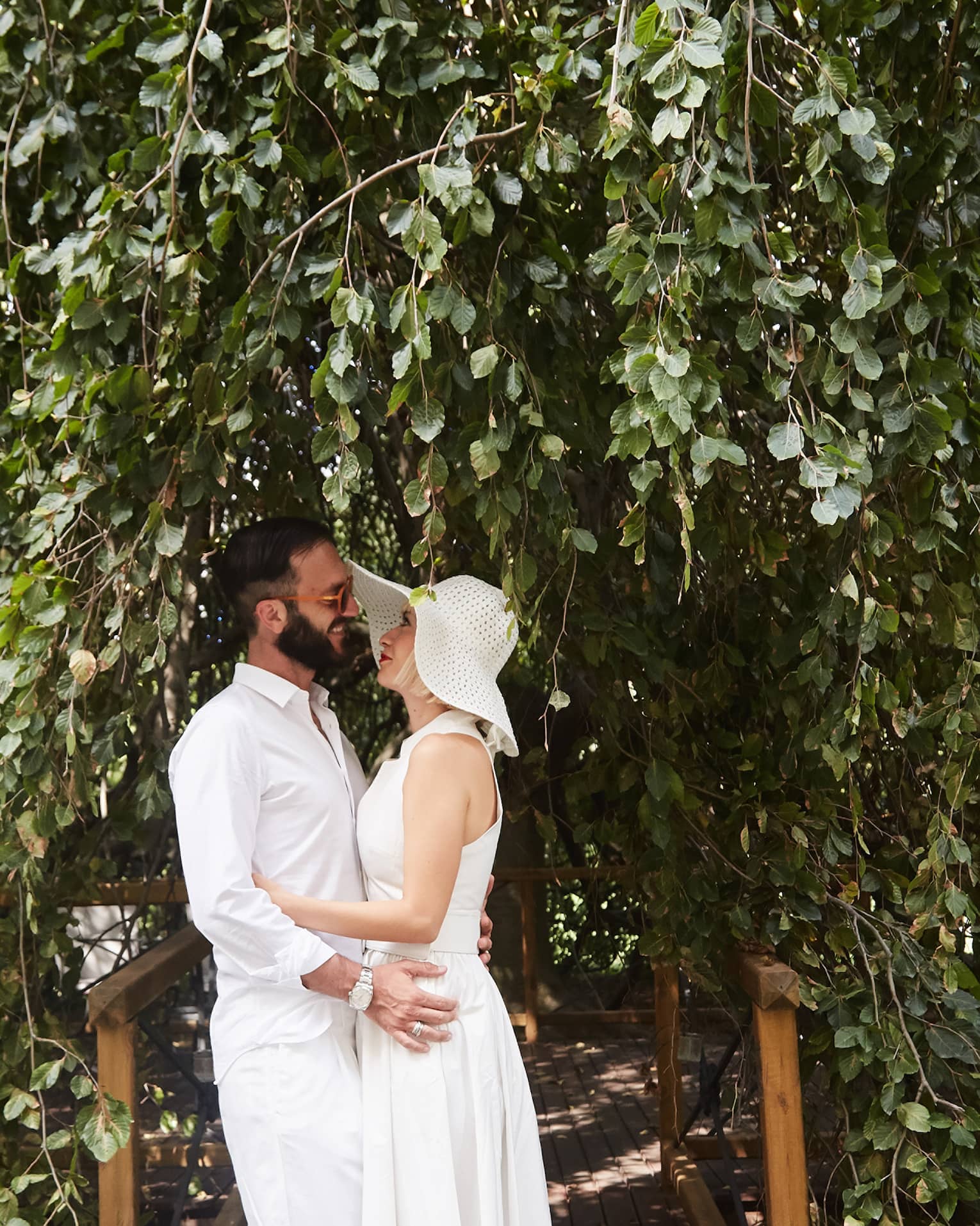 A couple embrace each other on a wooden path sheltered from the sun by long green branches cascading down around them.