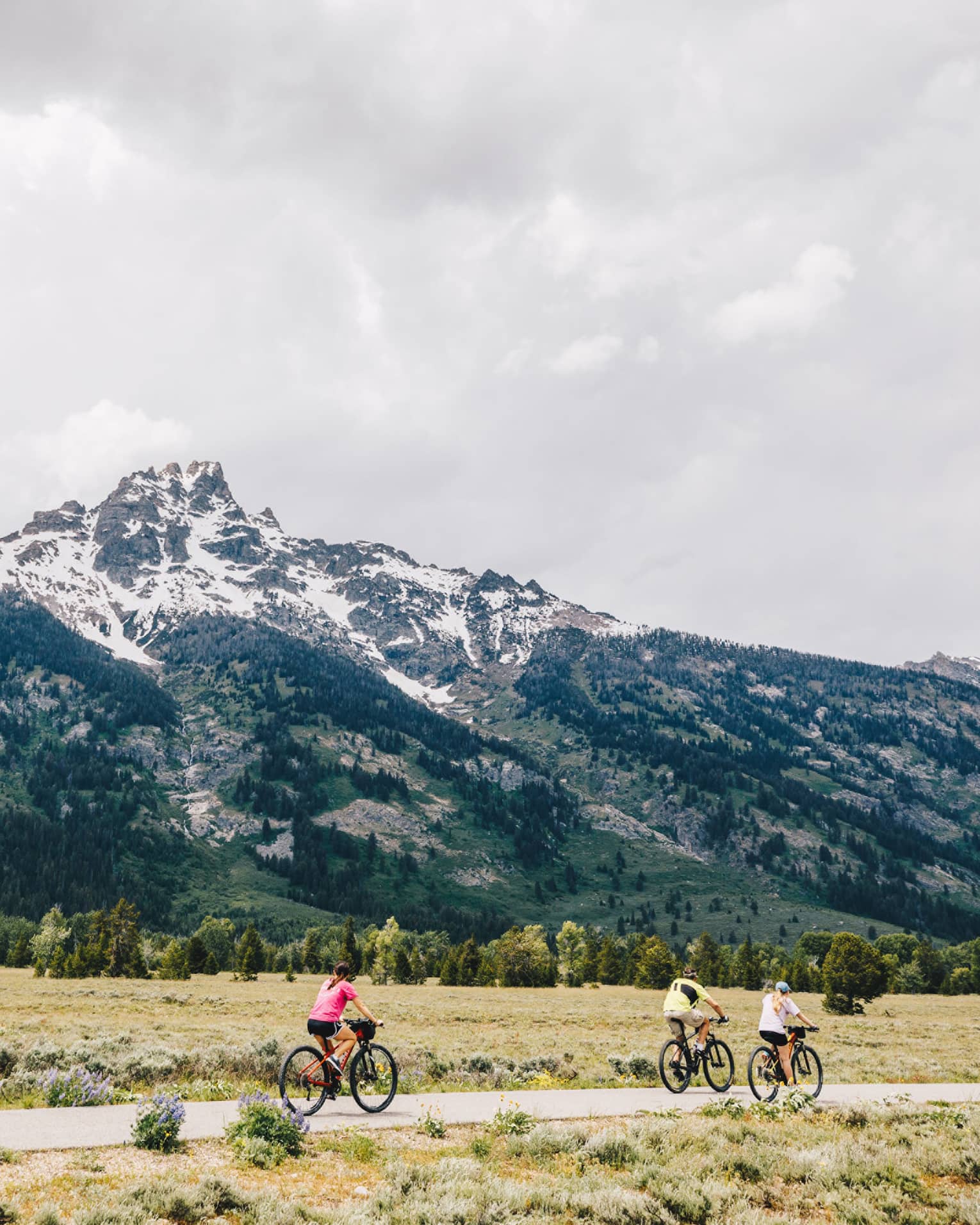 A man and two women cycle down a path on an overcast day with the mountains in the background