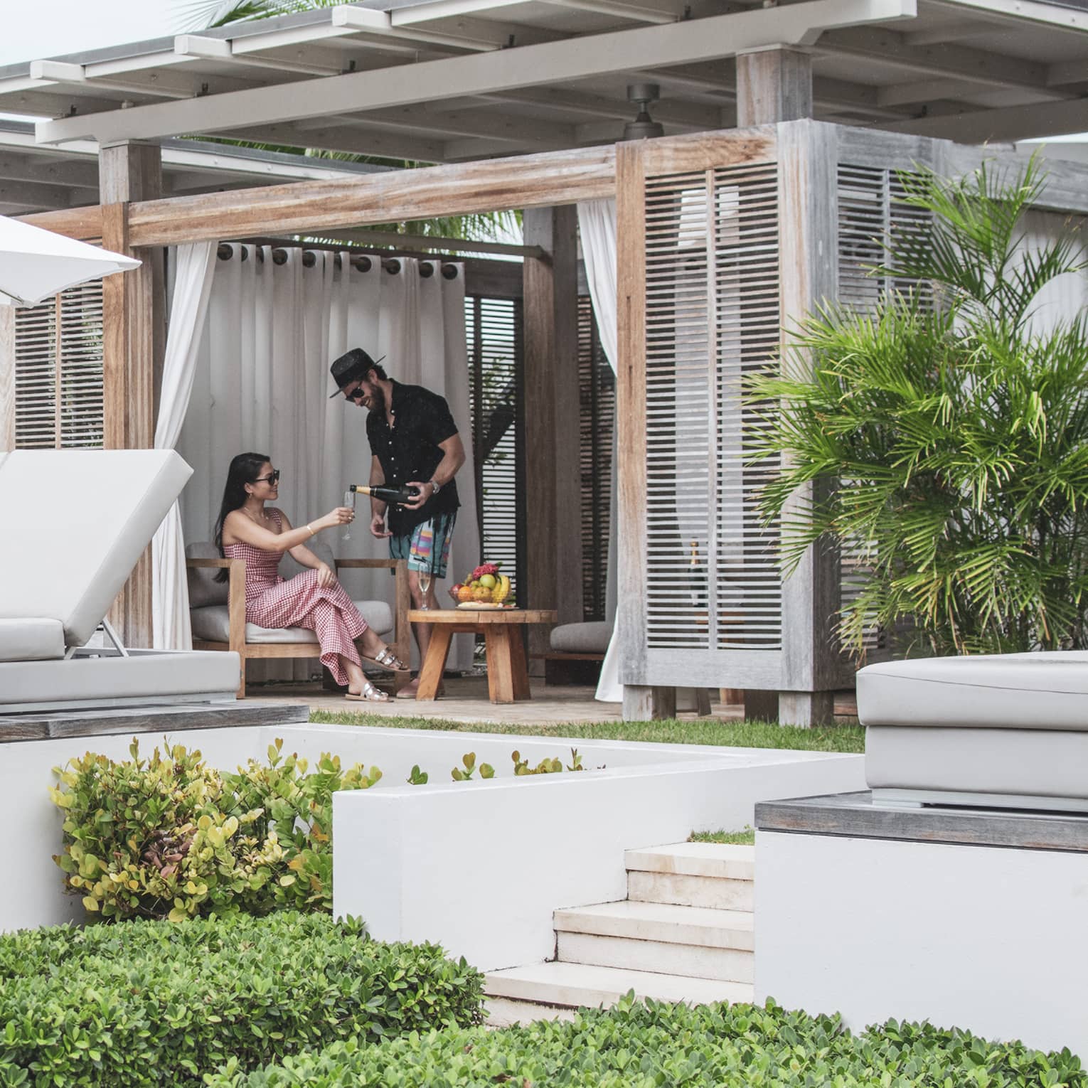 Two talk to each other in a covered pool cabana with a row of white daybeds and a row of grey lounge chairs and white umbrellas in front of them