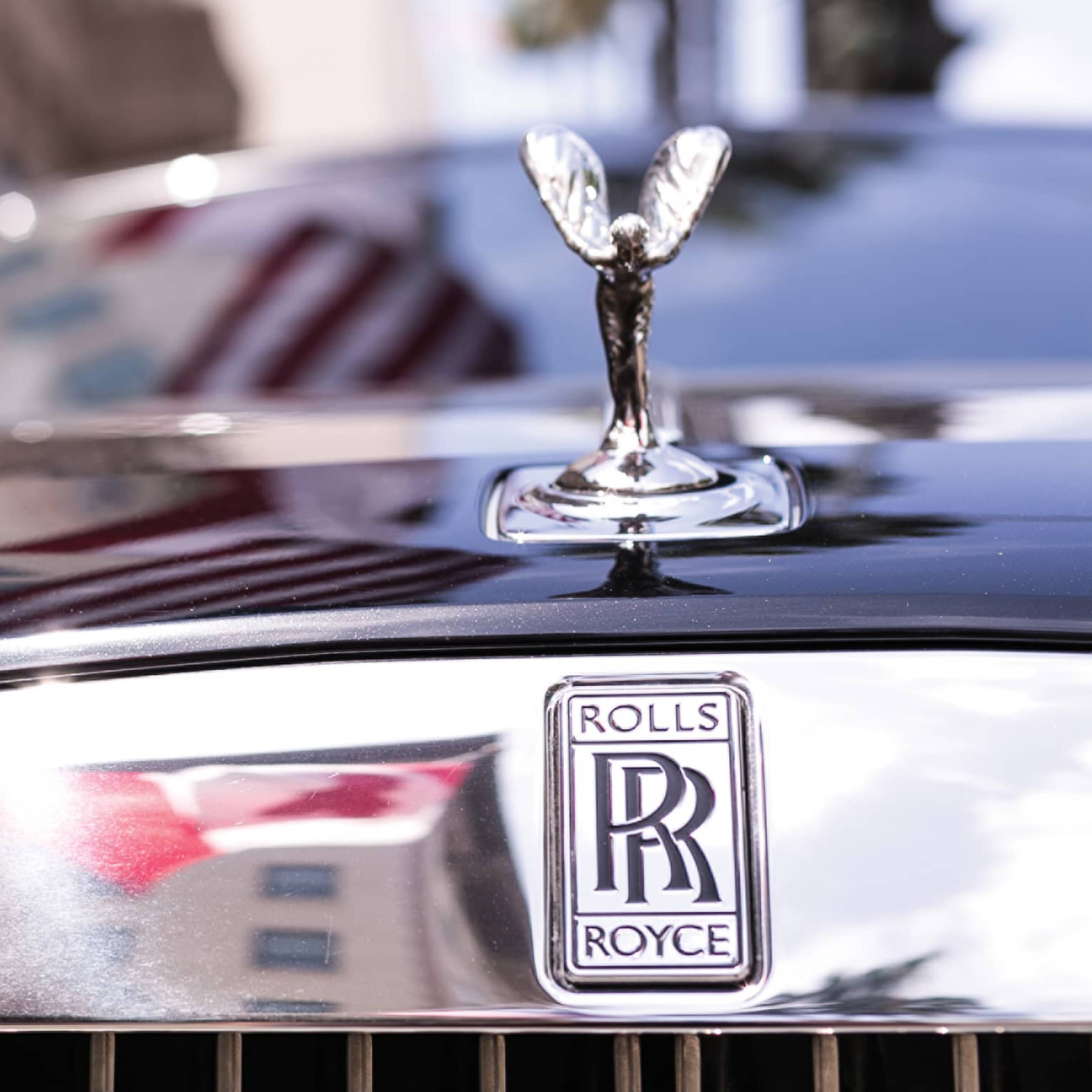 Close-up view of a winged silver ornament on the hood of black Rolls Royce, double-R logo on reflective chrome above grille.