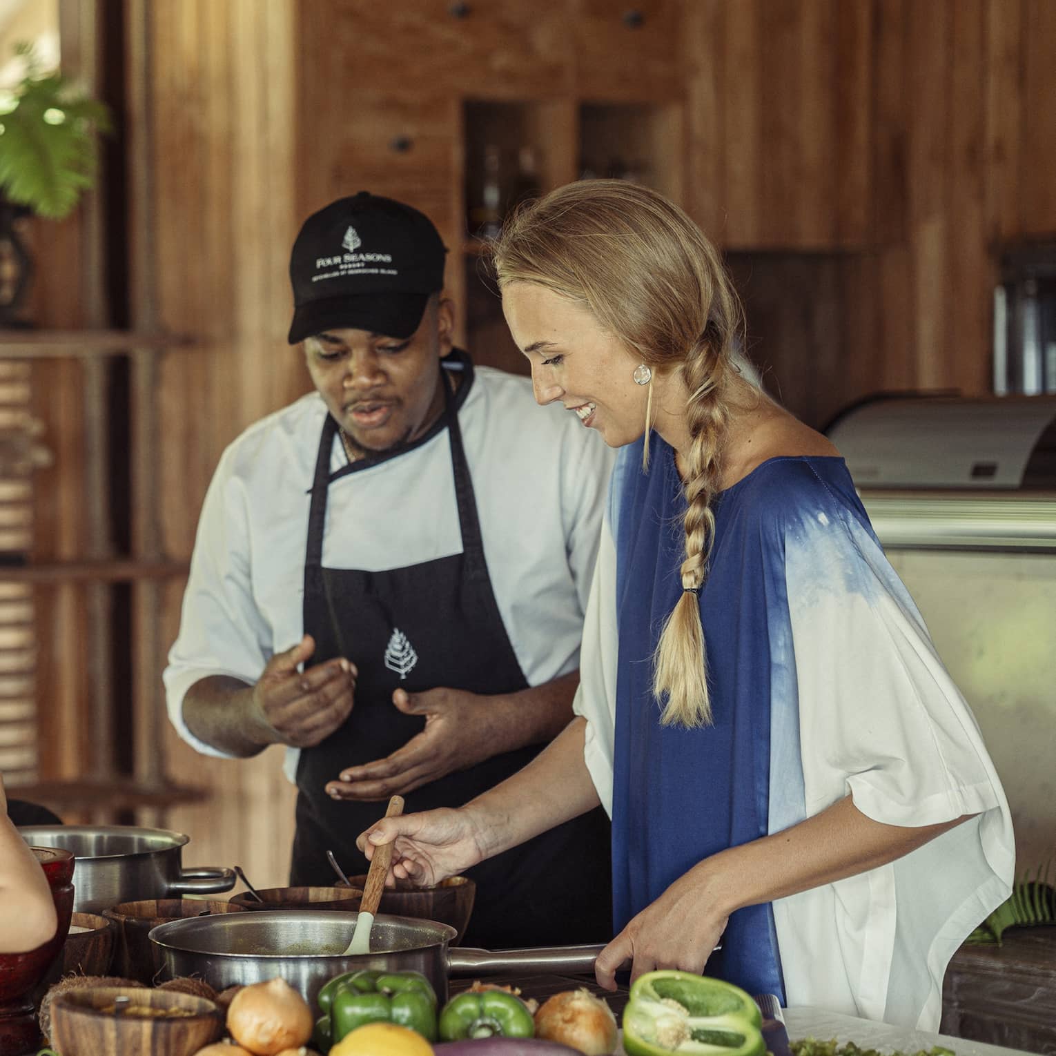 A chef instructs a guest stirring a pot, while a child uses a mortar and pestle across the counter as a third adult watches. 