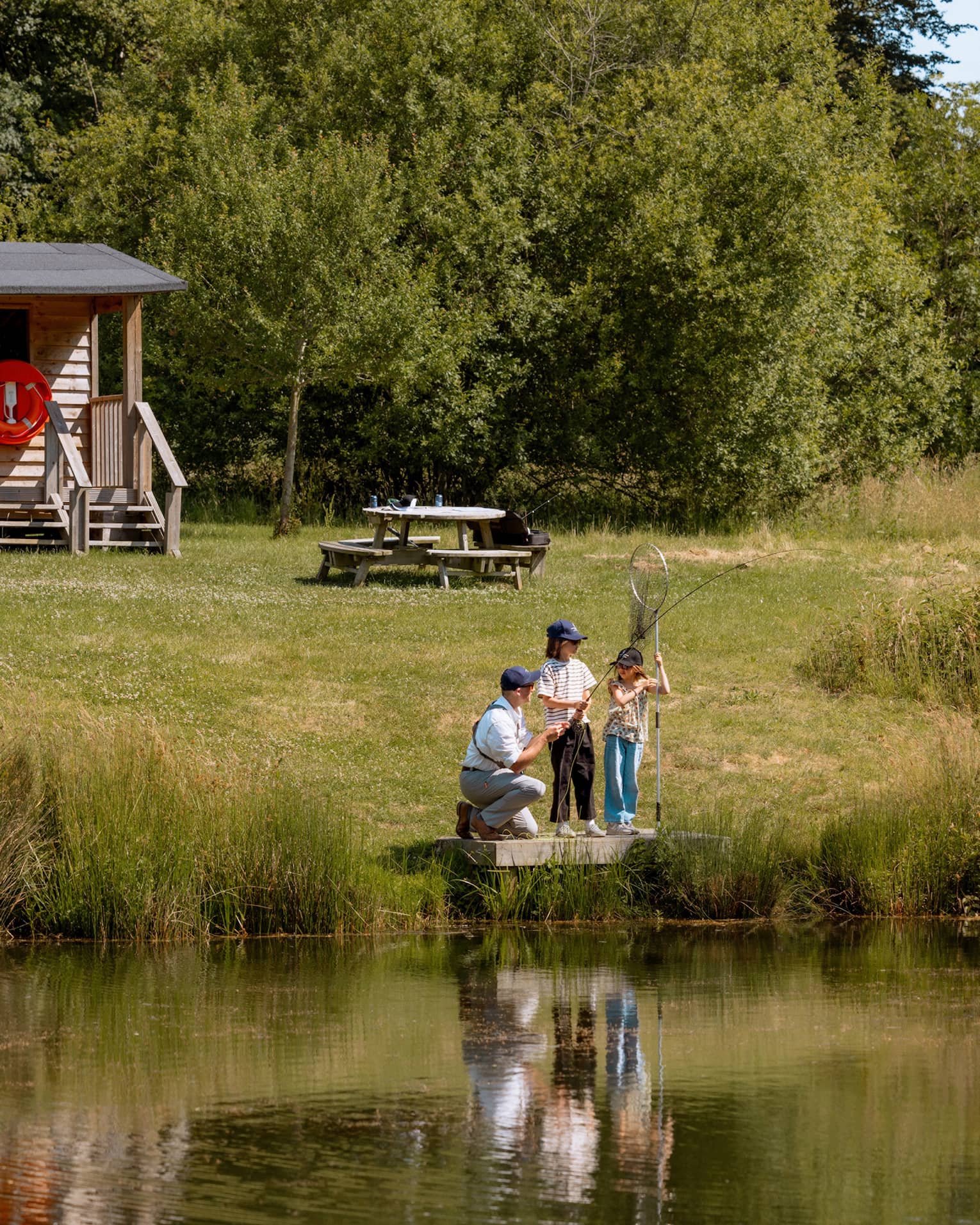 Three people fishing on small platform by lake with cabin-like building and picnic table in backdrop