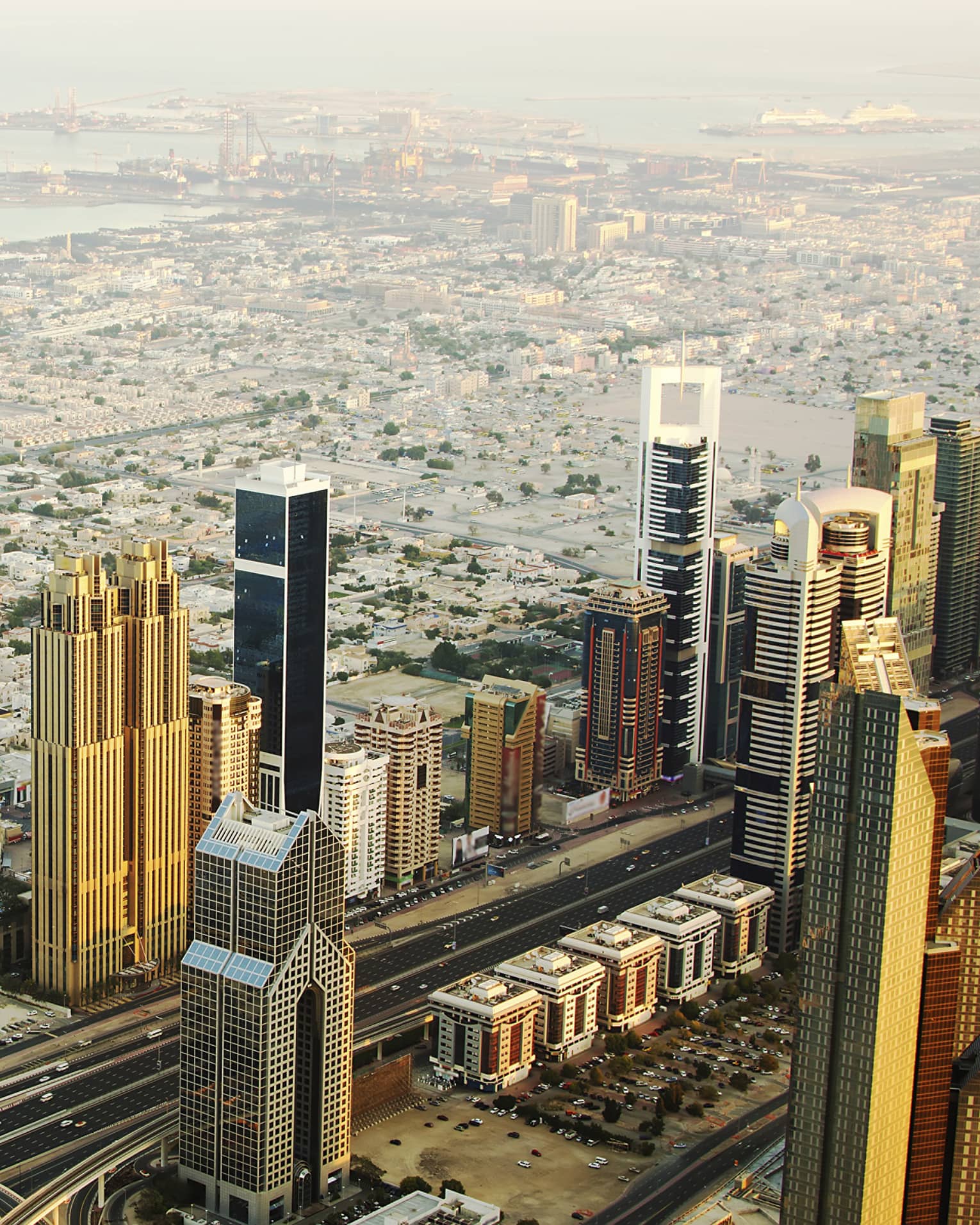 Aerial view of rows of golden towers reflecting sun against backdrop of low city grid receding into harbour under hazy sun.