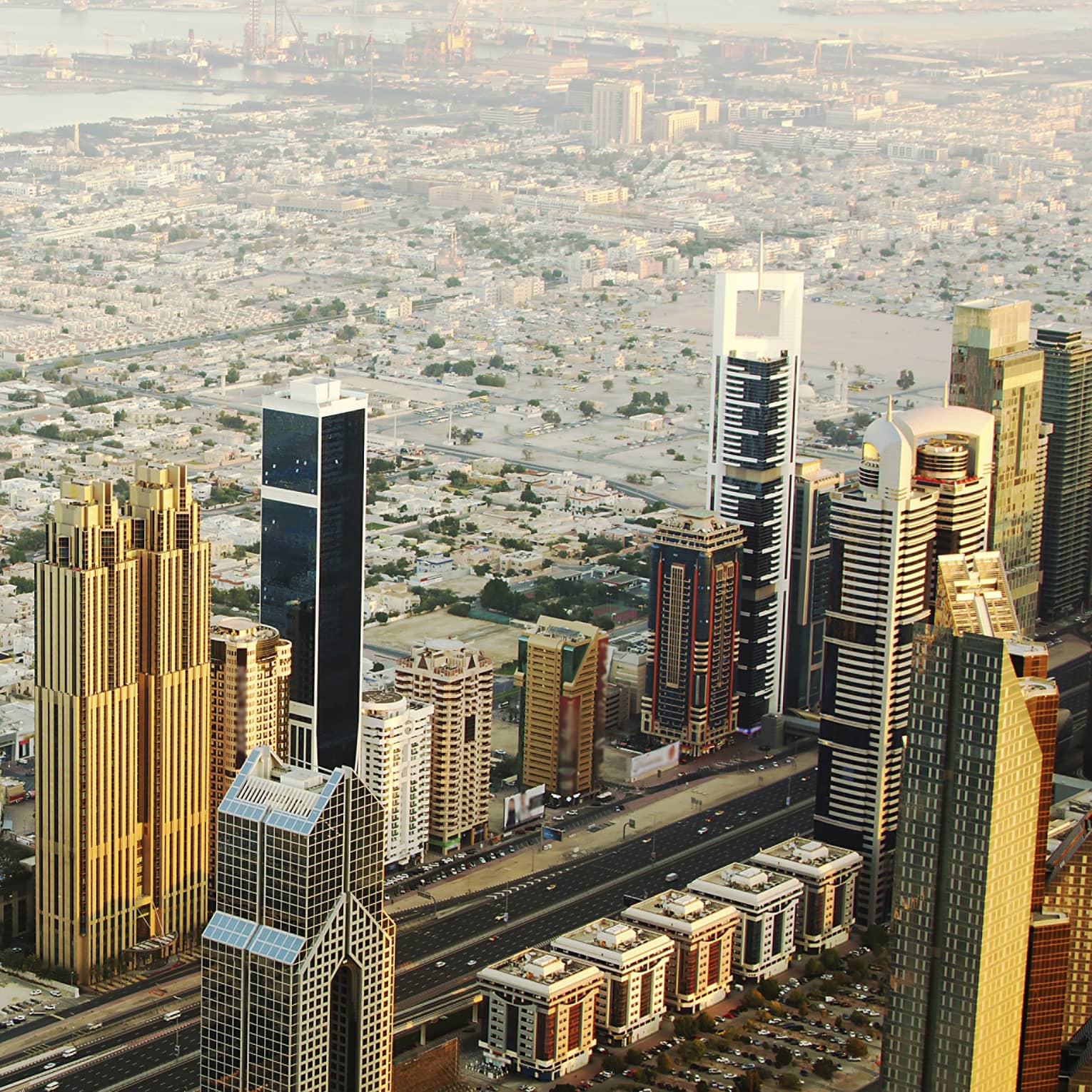 Aerial view of rows of golden towers reflecting sun against backdrop of low city grid receding into harbour under hazy sun.