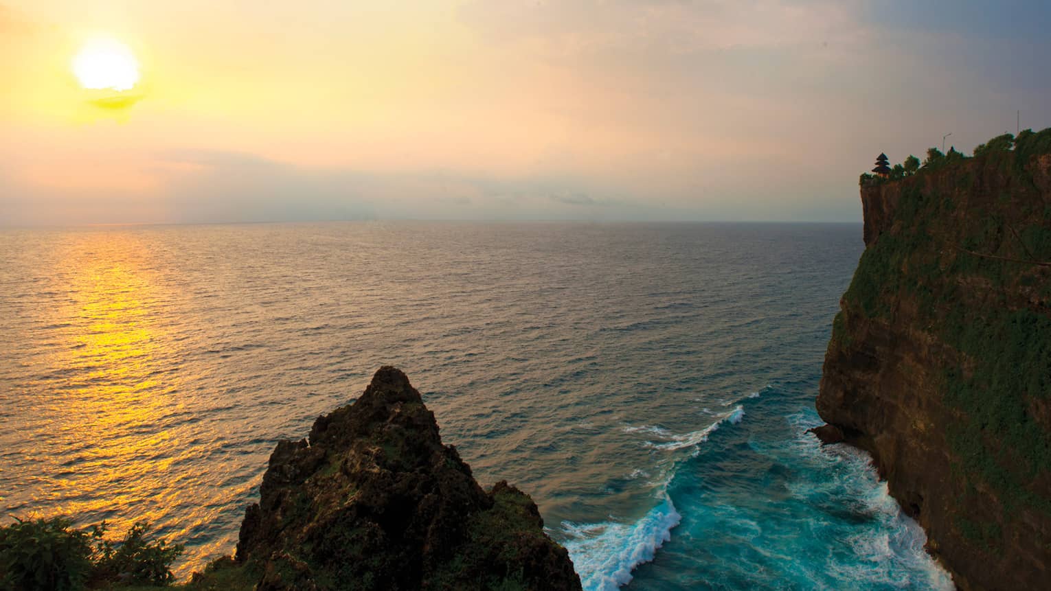 A hazy yellow sun reflects on a vast expanse of water with waves breaking against a tall, sheer cliff face dotted with trees.