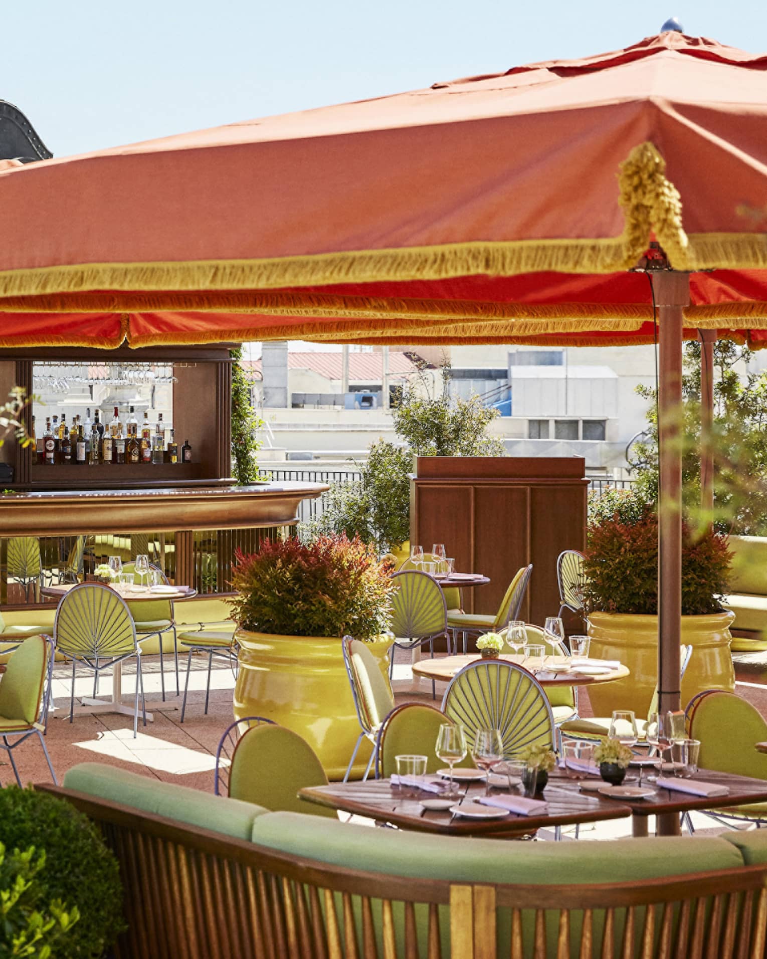 Outdoor terrace brasserie with green chairs and tables under an orange canopy, surrounded by plants, with statues and architectural details in the background