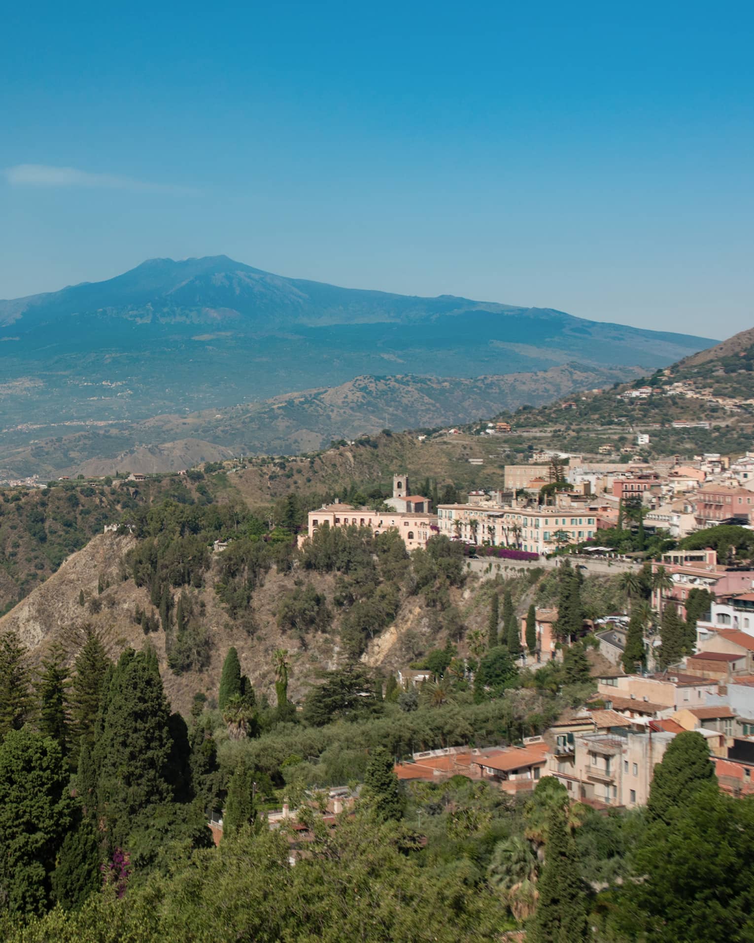Panoramic view of a coastal town nestled in the hills, overlooking the sea, with mountains in the background and greenery in the foreground.