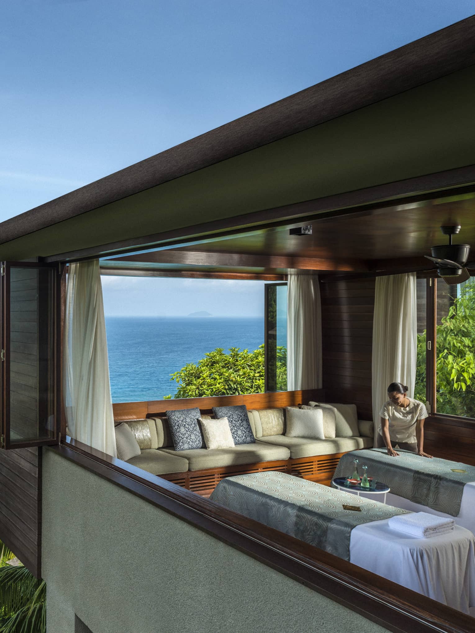 Woman smoothes sheet on side-by-side massage tables in spa room high above trees, ocean