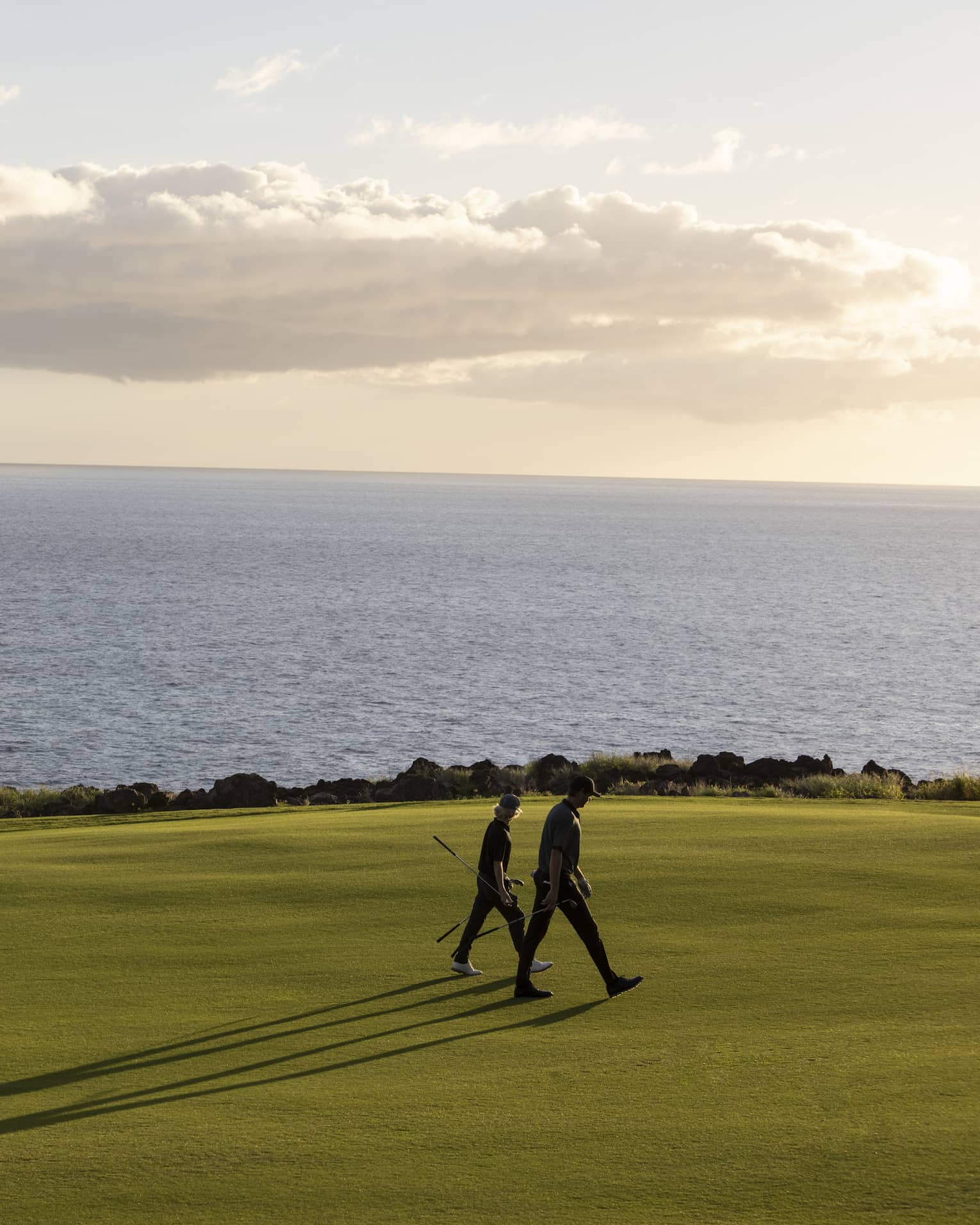 Two people with golf clubs, long shadows reaching out behind, walk a golf course set against calm ocean and fluffy clouds. 