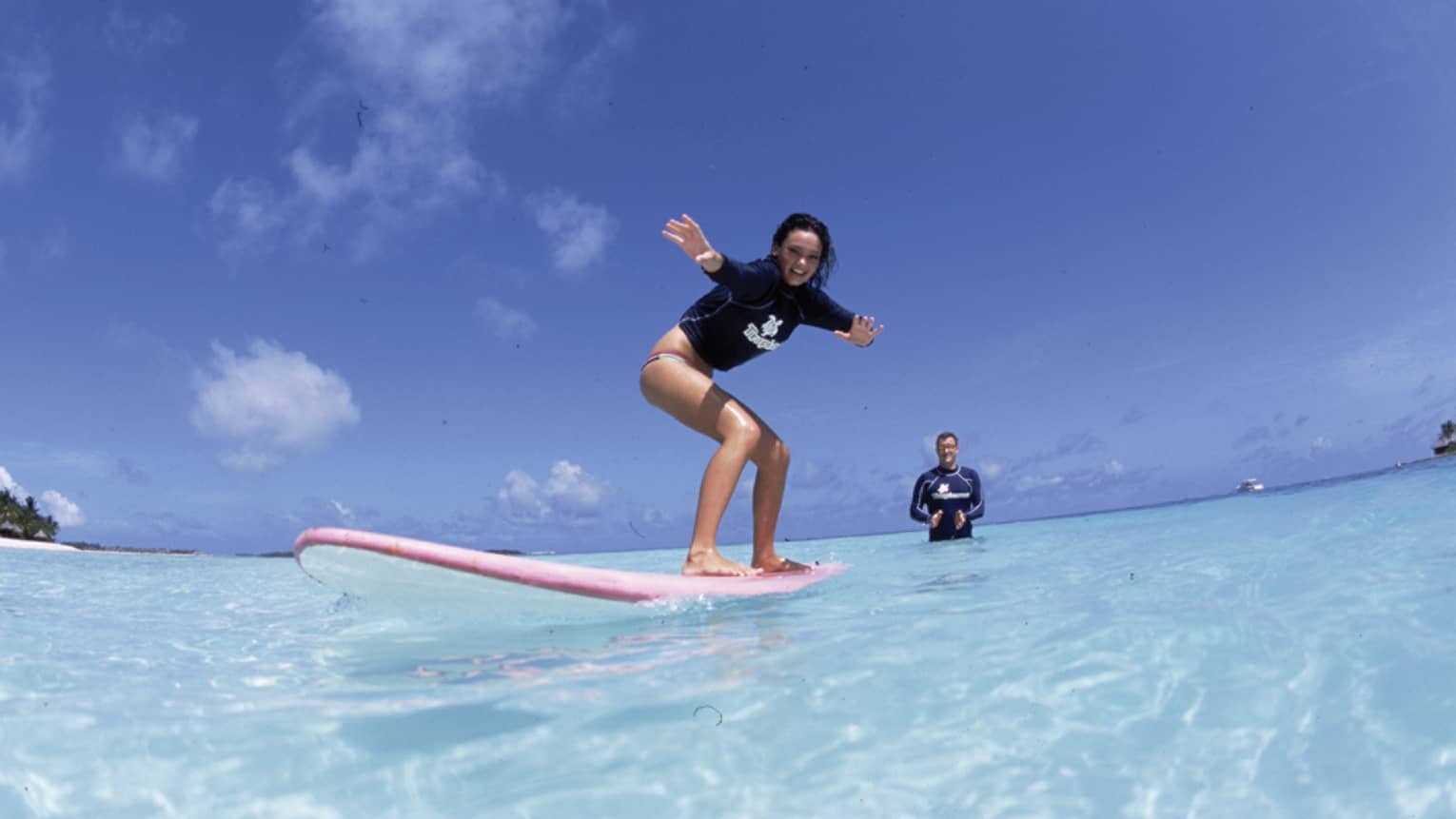 In the crystal-clear shimmering water is a woman learning to surf and an instructor standing in waist-deep water clapping.