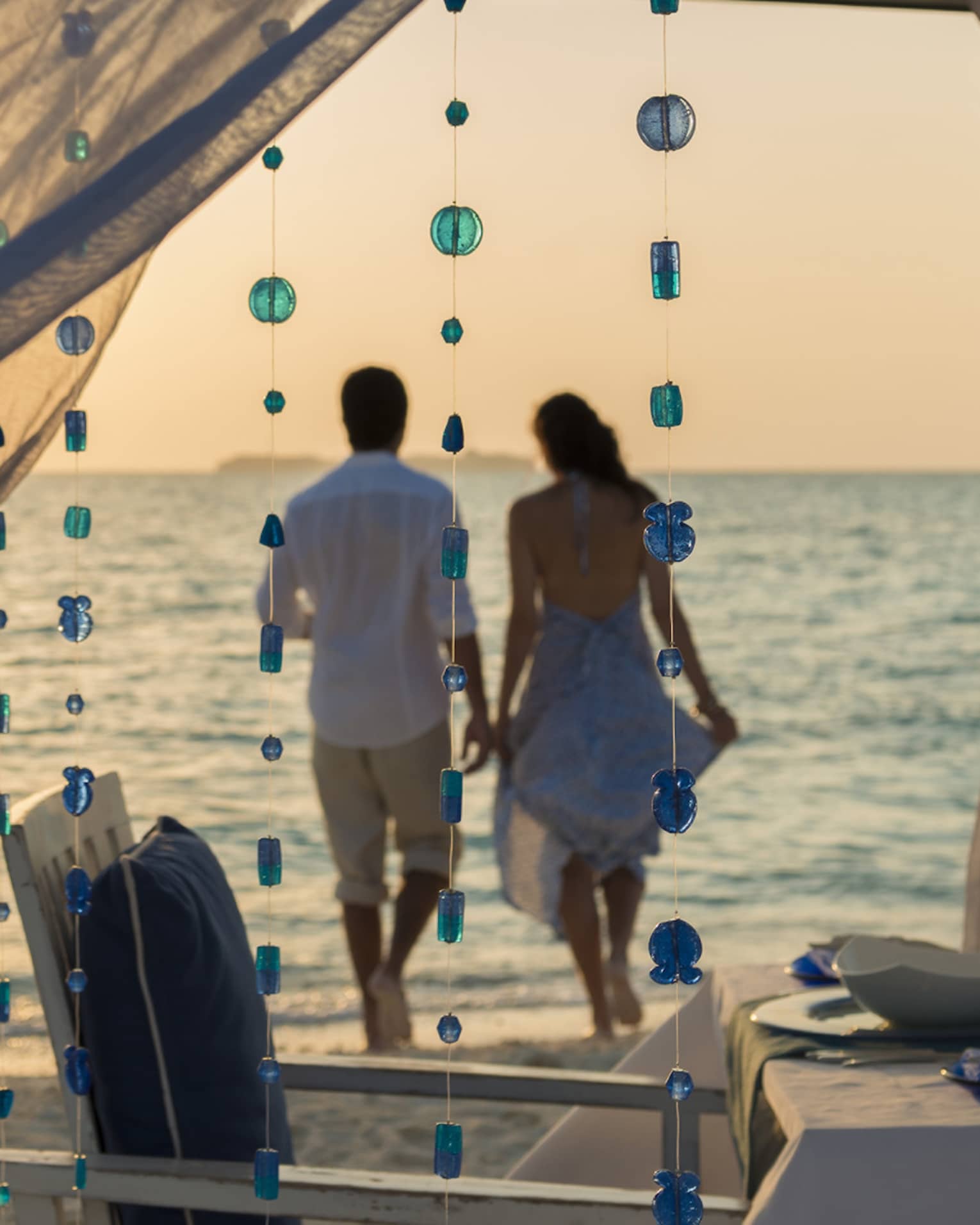 Sheer curtains blow in breeze, small blue beads hang from string, woman and man hold hands on beach in background