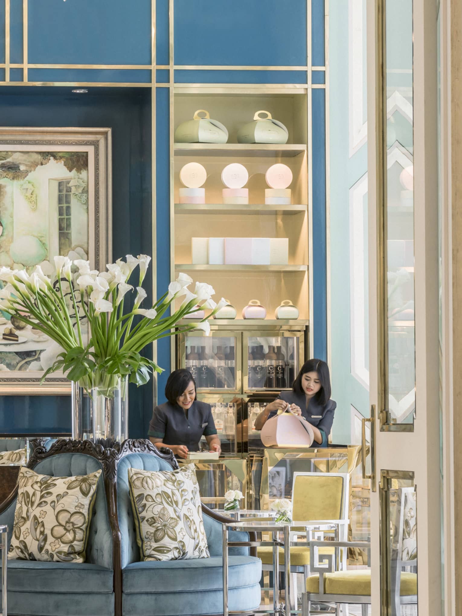 View inside La Patisserie doors with elegant blue and gold chairs, tall glass vase with white flowers, woman fastening pink box