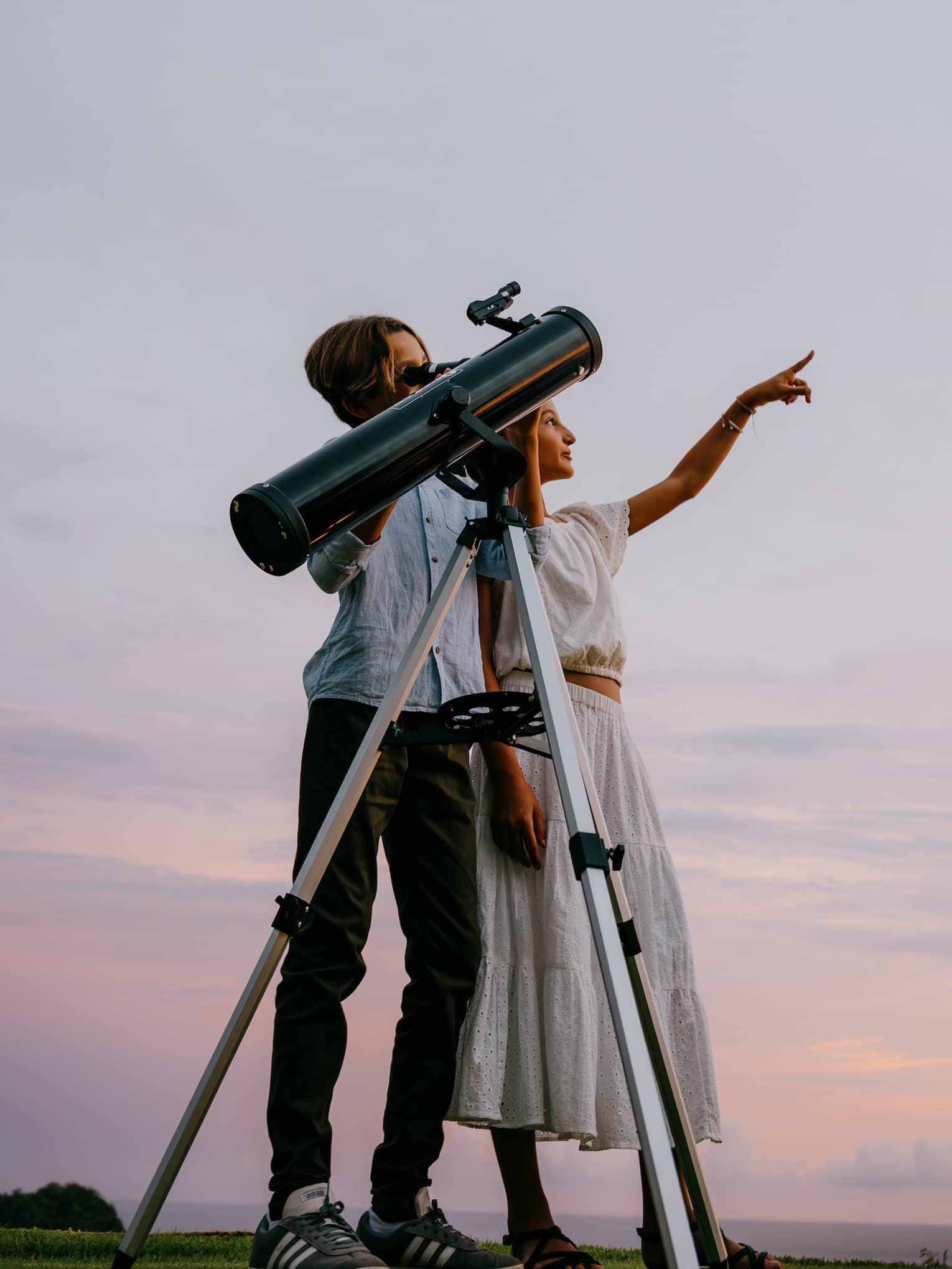 Two people stand next to a large telescope with an evening sky in the background. One person points toward the sky.