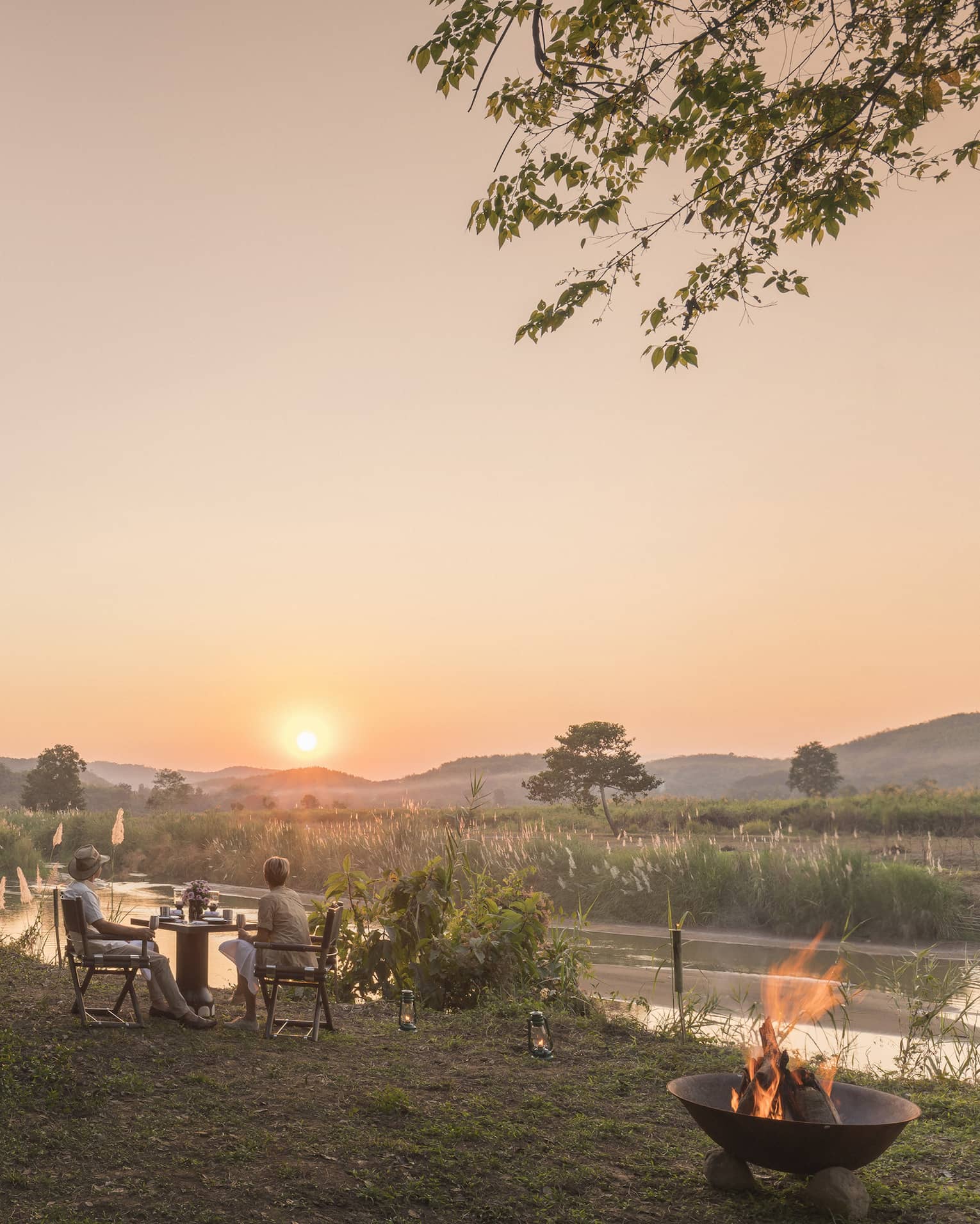 Outdoor riverside dining setup at sunset with a campfire and mountain views