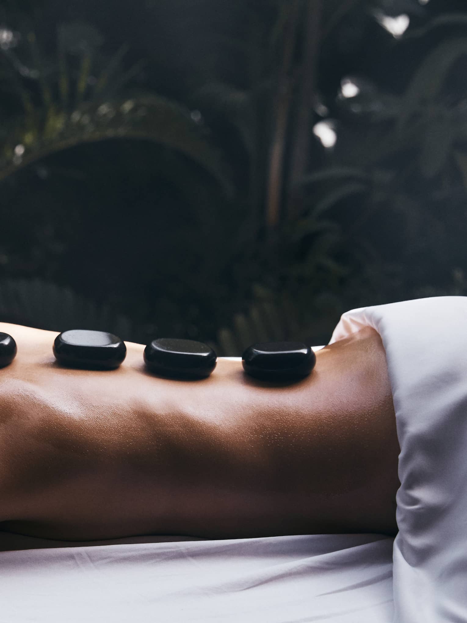 A guest on an outdoor massage table relaxes on their stomach with four glossy black warming stones on their back.