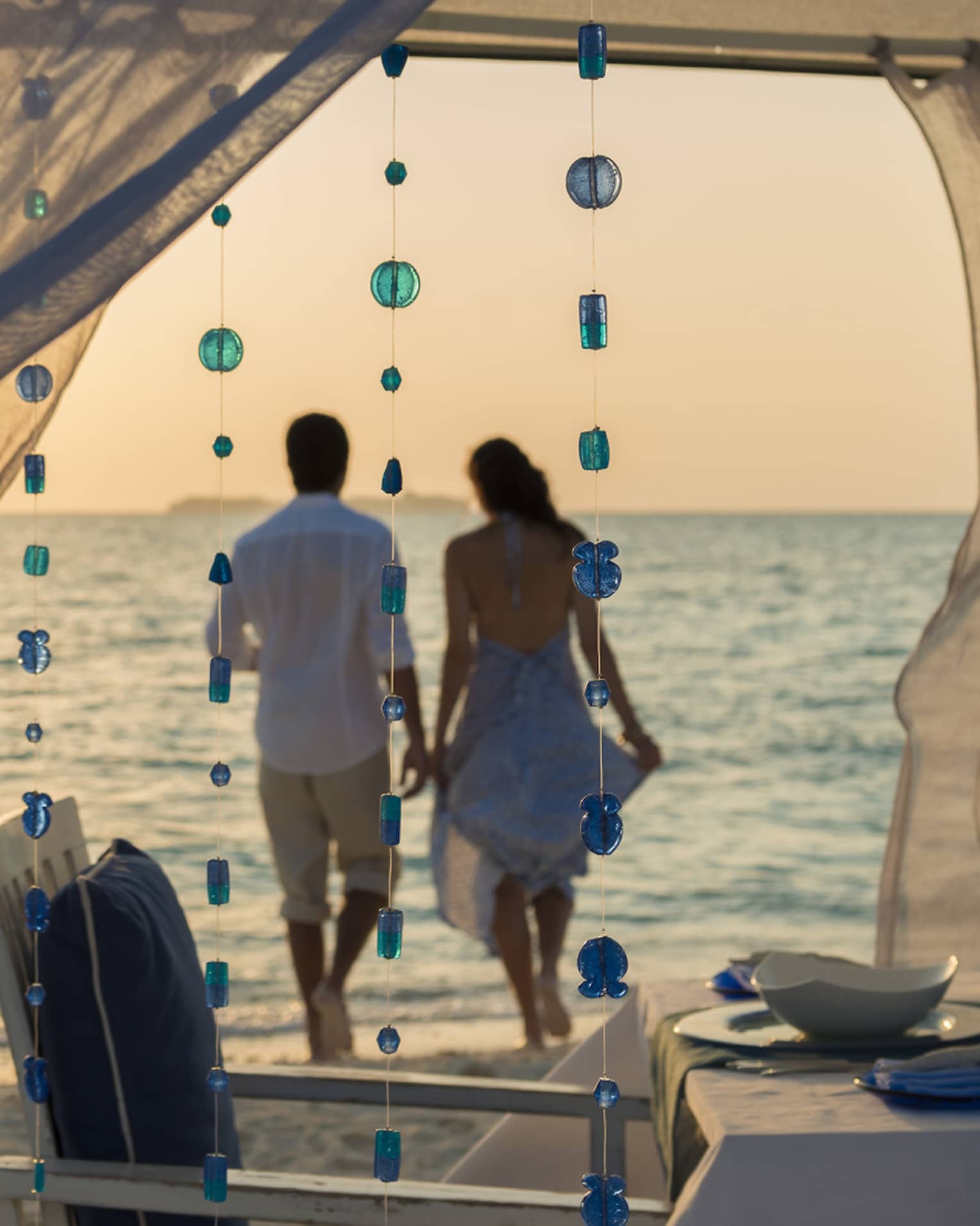 Sheer curtains blow in breeze, small blue beads hang from string, woman and man hold hands on beach in background