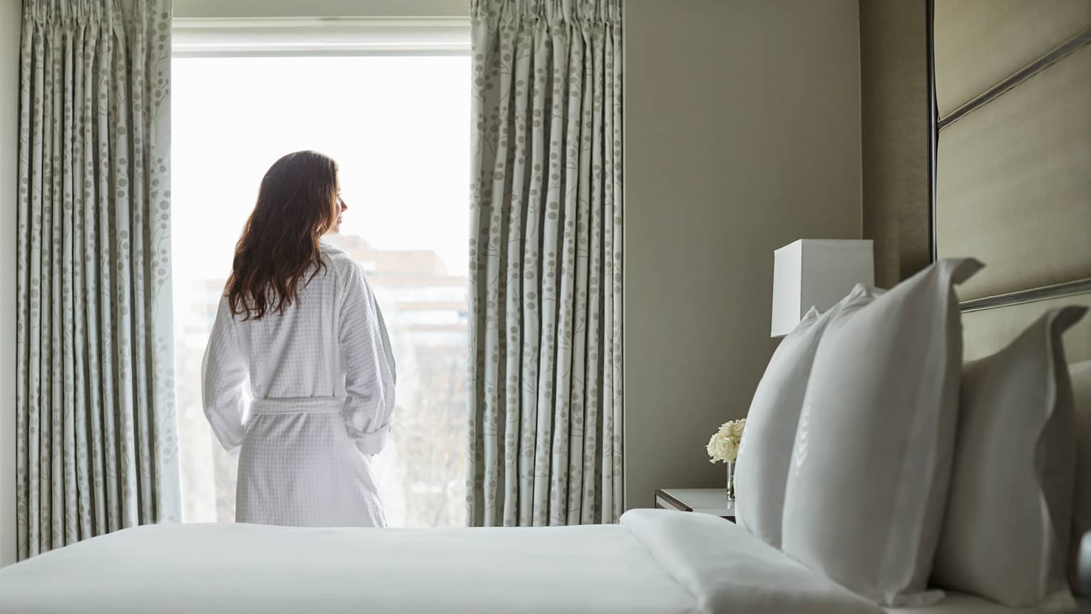 Back of woman in white bathrobe stands at sunny window in front of bed