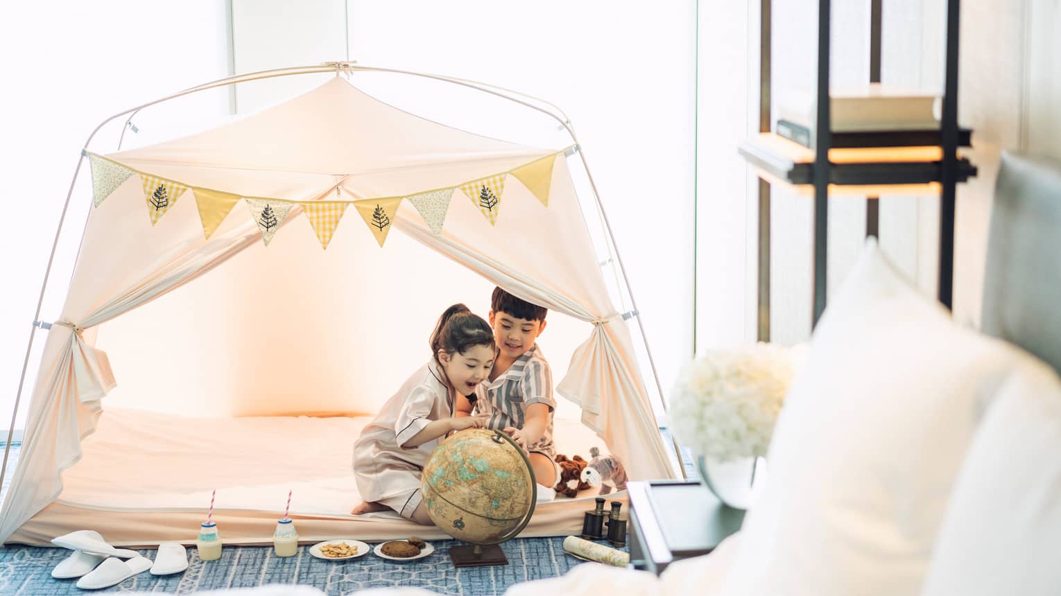 Two children wearing white bathrobes play in white tent in hotel room by bed