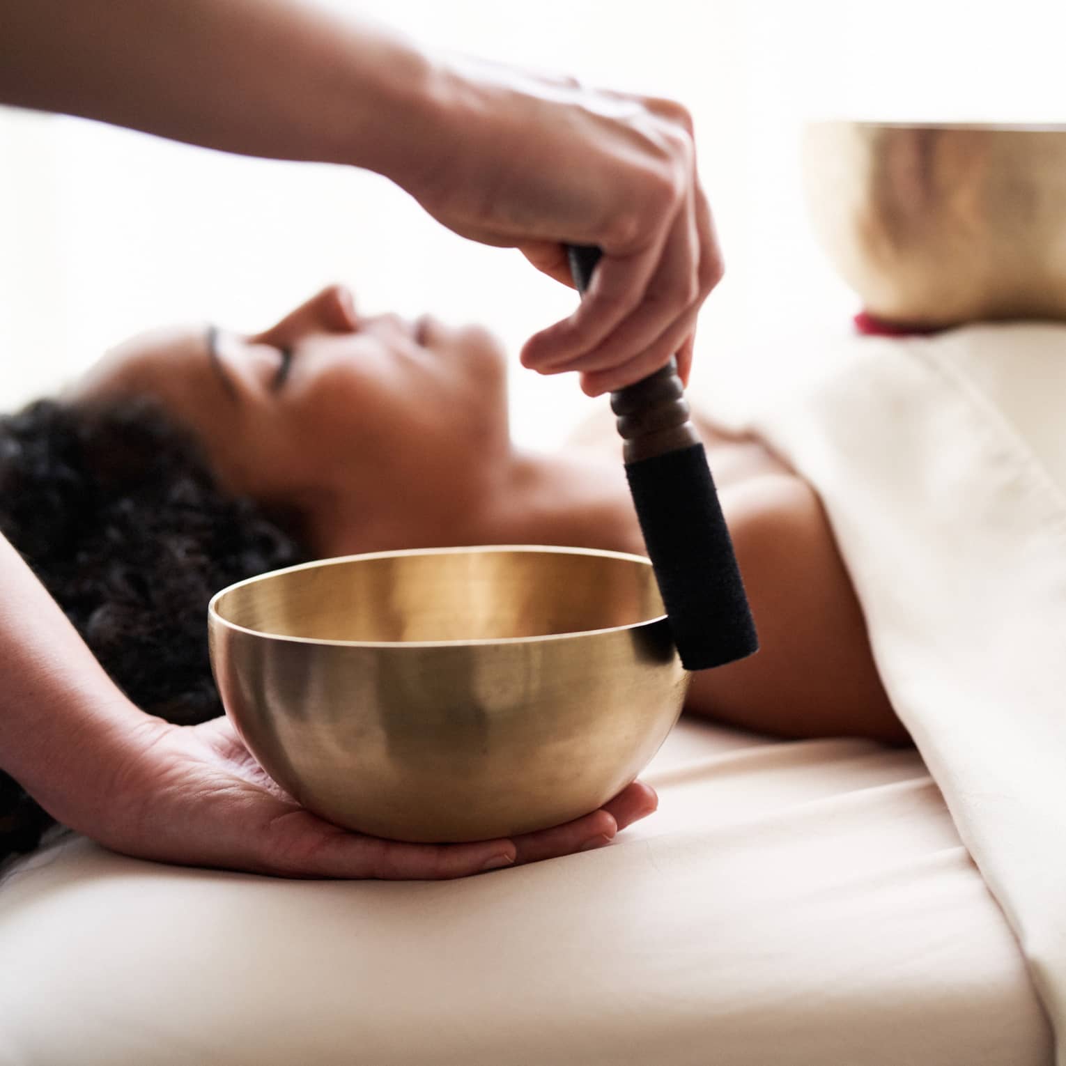 Woman with eyes closed lies on massage table in as vibrational sound therapy is performed with a Tibetan singing bowl