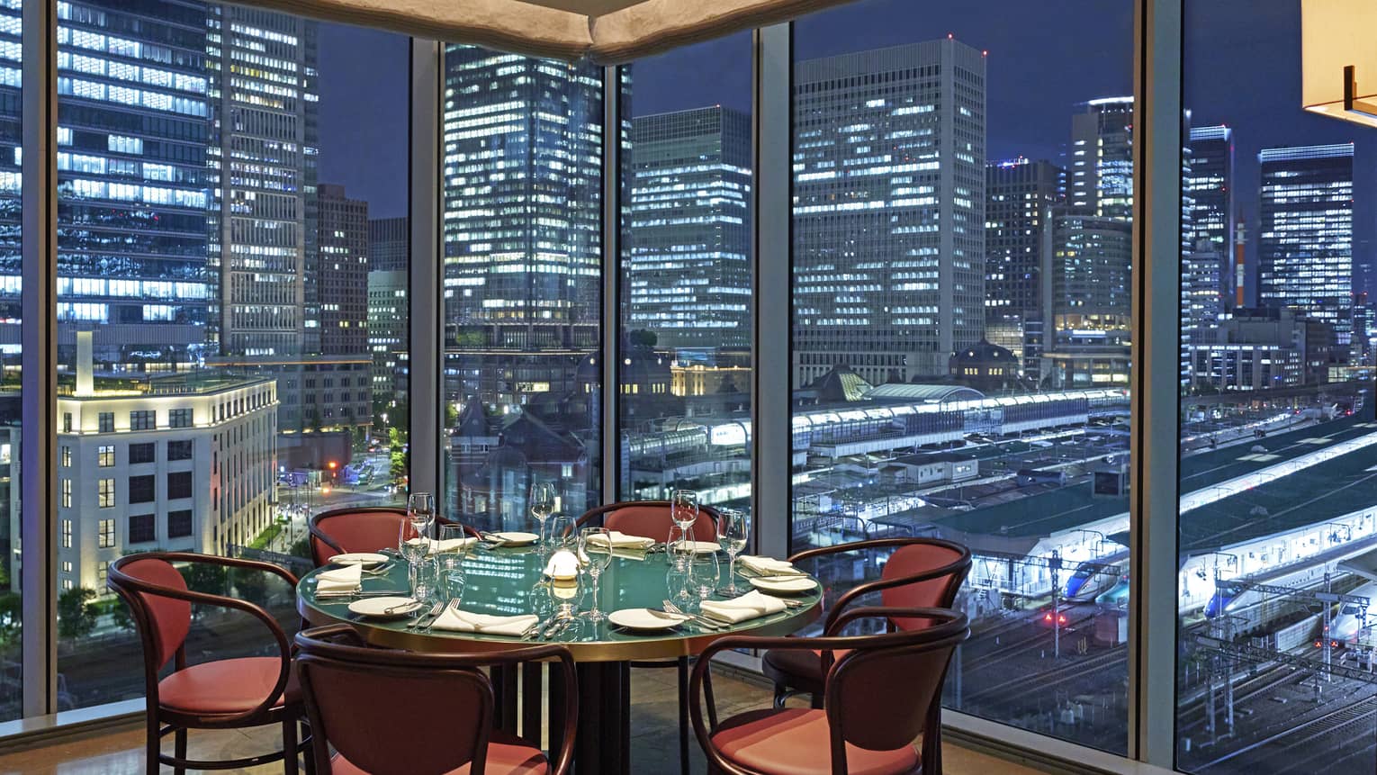 Luxury boutique hotel dining setup with a round table elegantly set for guests, offering a panoramic city view at night through tall glass windows