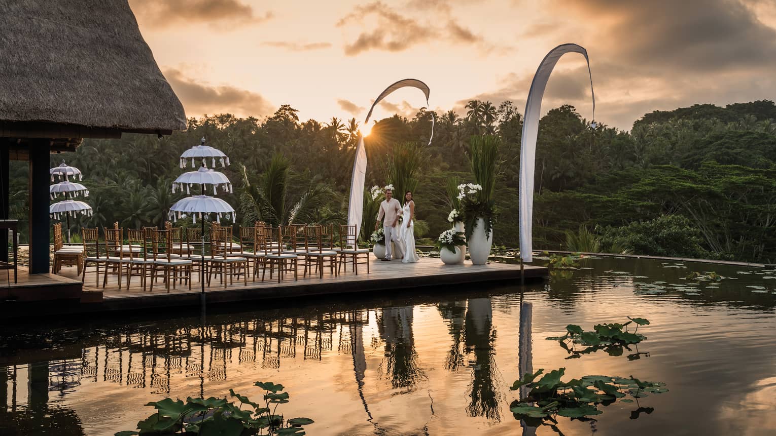 Bride and groom stand by wedding ceremony chairs at edge of patio, outdoor pool reflecting sunset