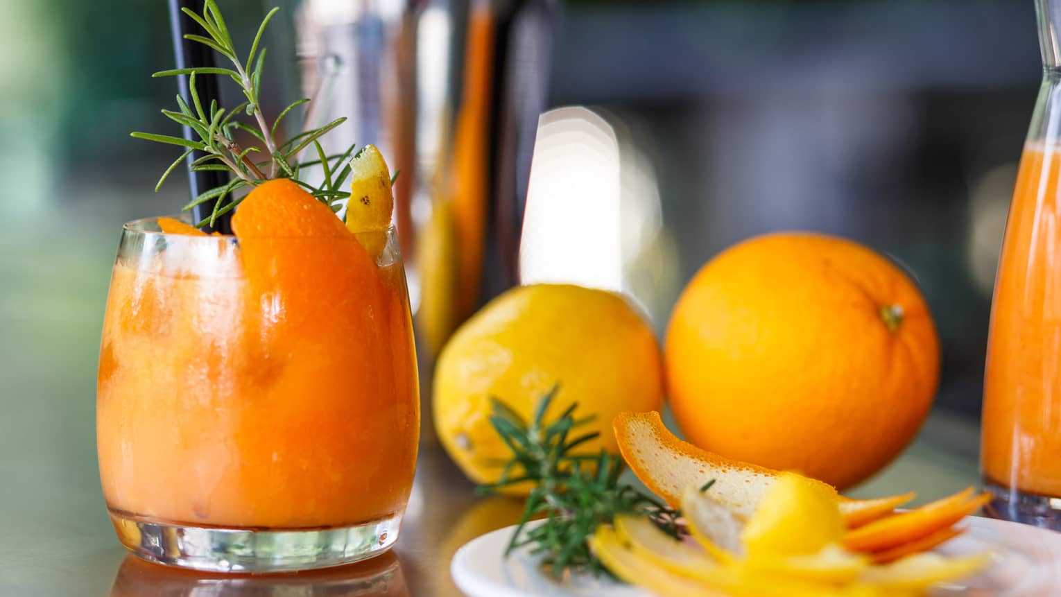 Bright orange cocktail garnished with lemon and orange rinds, rosemary sprig, whole oranges and peels on side