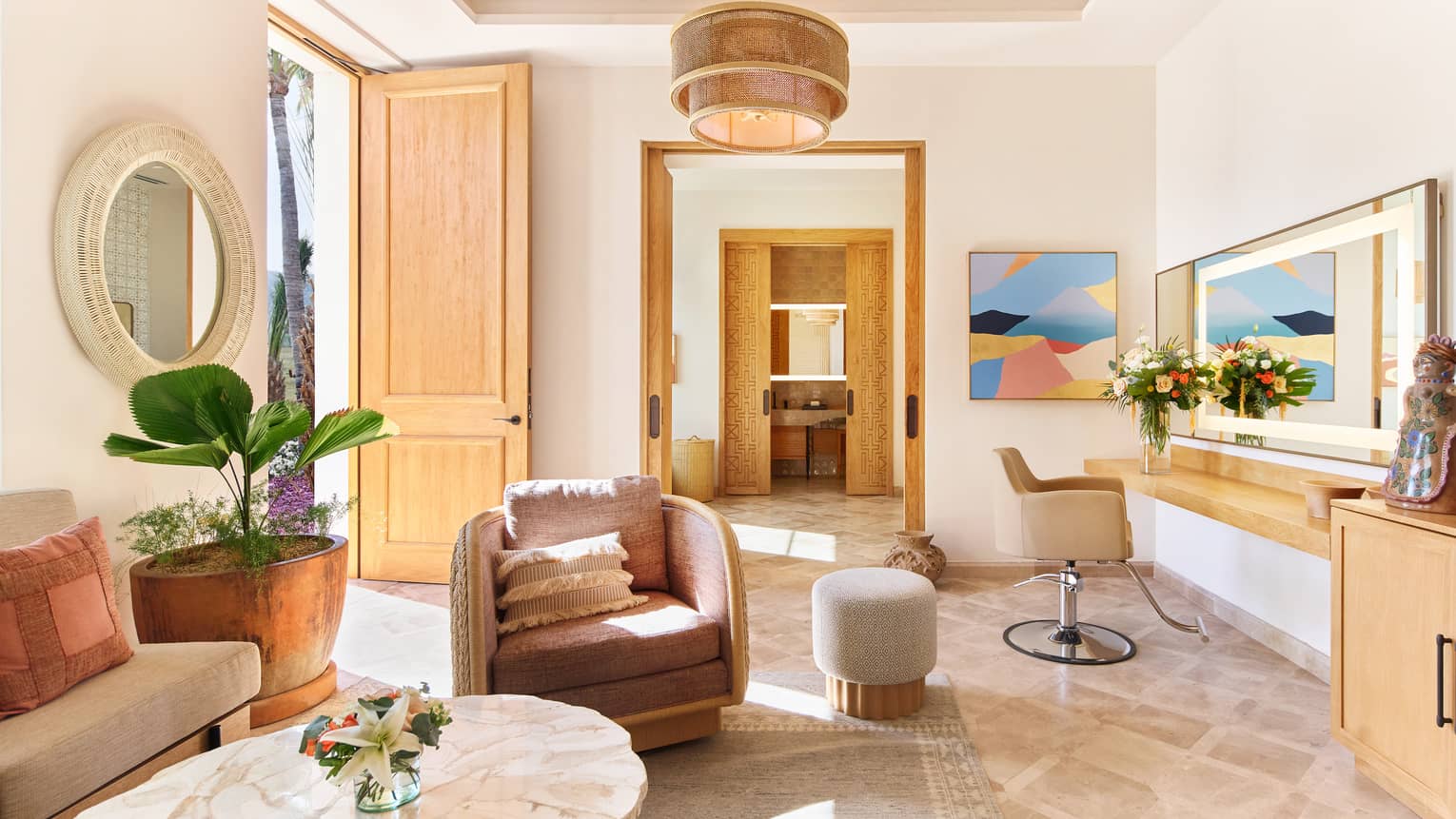 A spacious room filled with natural light, warm neutral decor, comfortable upholstered seating, artwork on the walls, floral arrangements and potted plants, and coffee and end tables