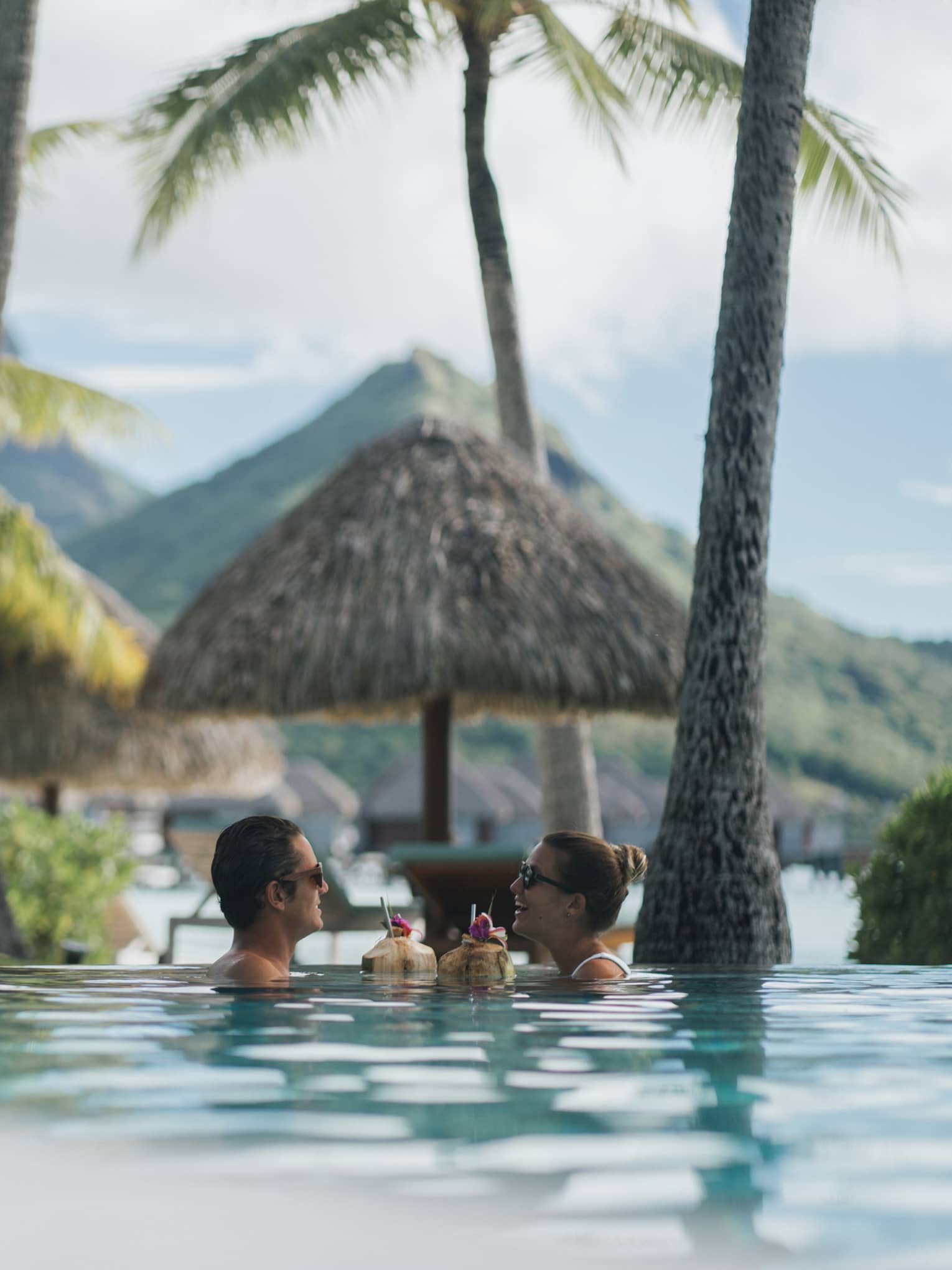 Two guests sip coconut drinks while floating in a pool surrounded by palm trees