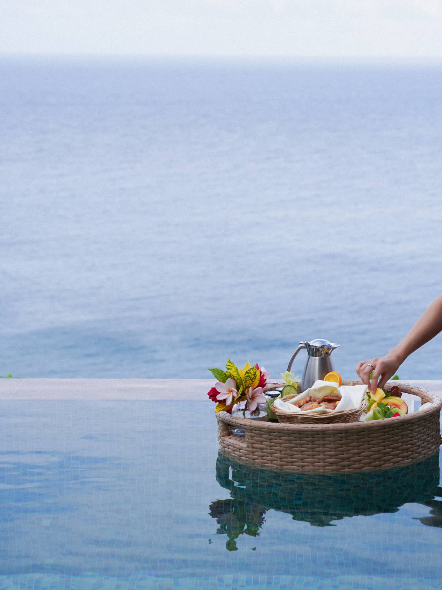 A hand delicately reaches into a floating basket of food, fruits, flowers and a carafe in a pristine blue ocean-view pool.