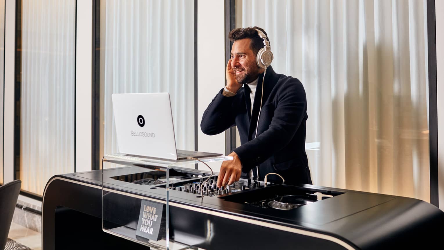 A DJ performing in a white room with closed windows behind him.